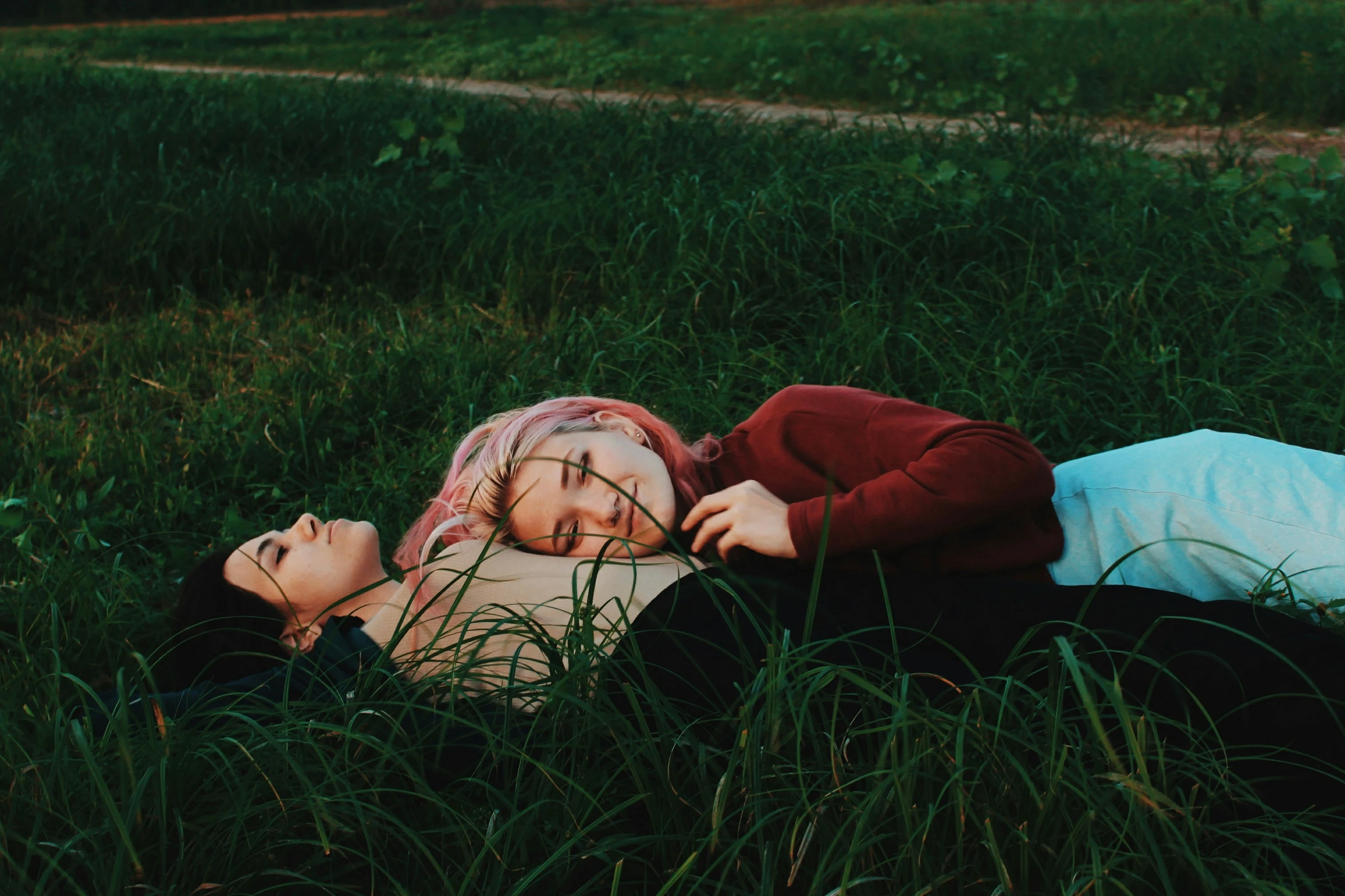 Two queer partners lie together in tall green grass at dusk, one resting gently on the other’s chest in a moment of quiet intimacy and emotional safety. The soft evening light and natural setting evoke themes of grounding, vulnerability, and authentic connection within LGBTQ+ relationships. This image reflects the heart of spiritually queer relationship therapy—creating space beyond heteronormative expectations to explore love consciously and intentionally. The couple’s relaxed posture and physical closeness symbolize trust, emotional presence, and the deep work of learning how to both give and receive love. Surrounded by nature, the scene mirrors the therapeutic journey: slowing down, softening defenses, releasing right-and-wrong narratives, and cultivating relational growth rooted in spiritual awareness, acceptance, and mutual care. Ideal for content focused on queer couples counseling, LGBTQ+ relationship healing, minority stress, emotional intimacy, and transformative love.