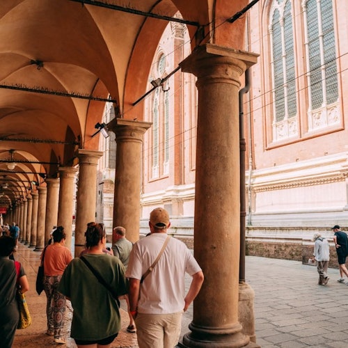 אנשים הולכים תחת קשתות עמודים גבוהים ליד מבנה לבנים היסטורי עם חלונות גדולים.