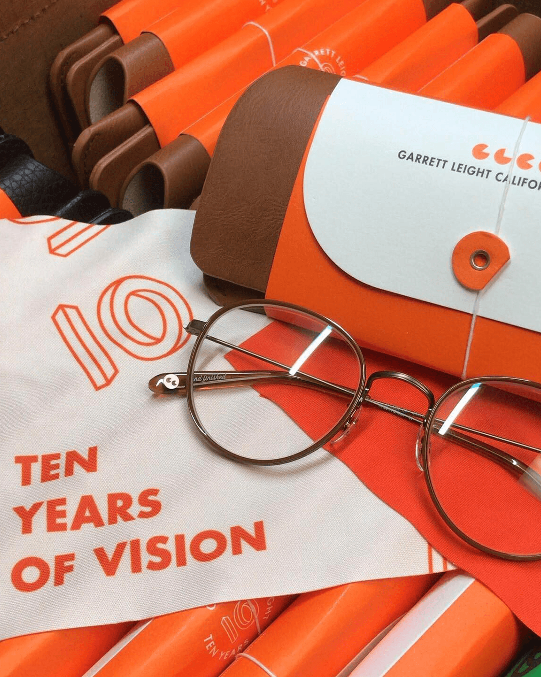 Close-up of Garrett Leight 10-year anniversary campaign collateral: stacked lens cases, glasses, and orange branded packaging with the "Ten Years of Vision" campaign typography