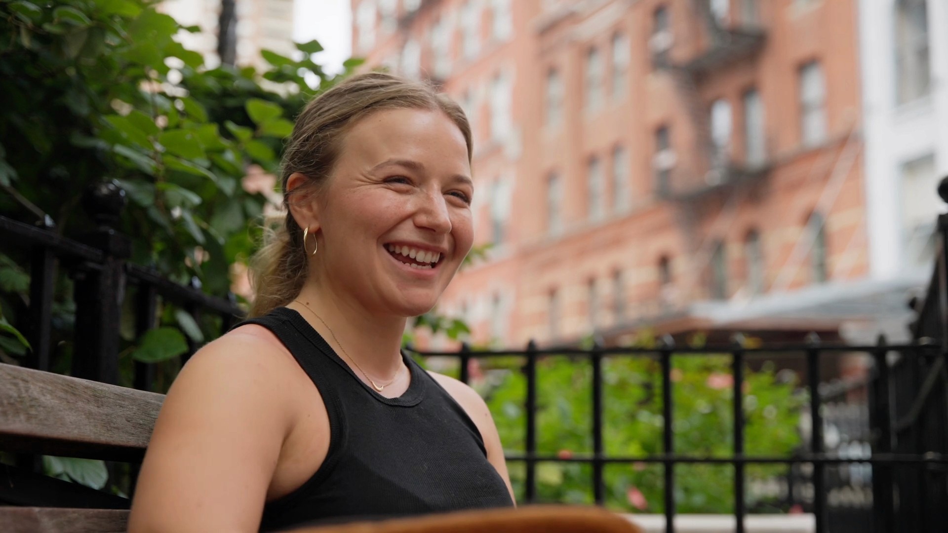 A person with blonde hair smiling at the camera, outdoors with buildings in the background