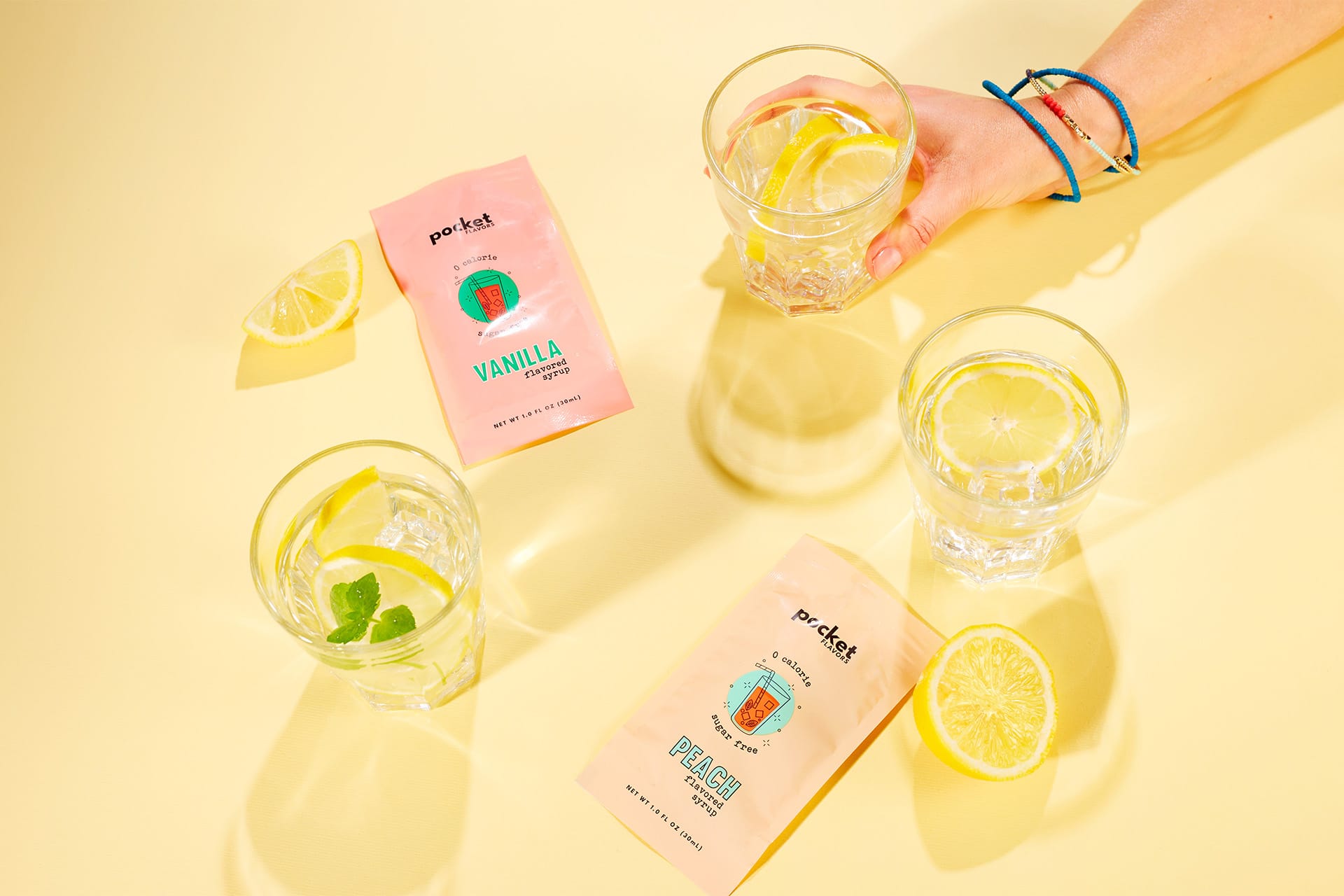 Ręka nosząca bransoletki trzyma szklankę z wodą cytrynową otoczoną saszetkami mieszanki napojów Pocket Flavors o smaku wanilii i brzoskwini na jasnop Yellow tle, z plasterkami cytryny rozsypanymi wokół.