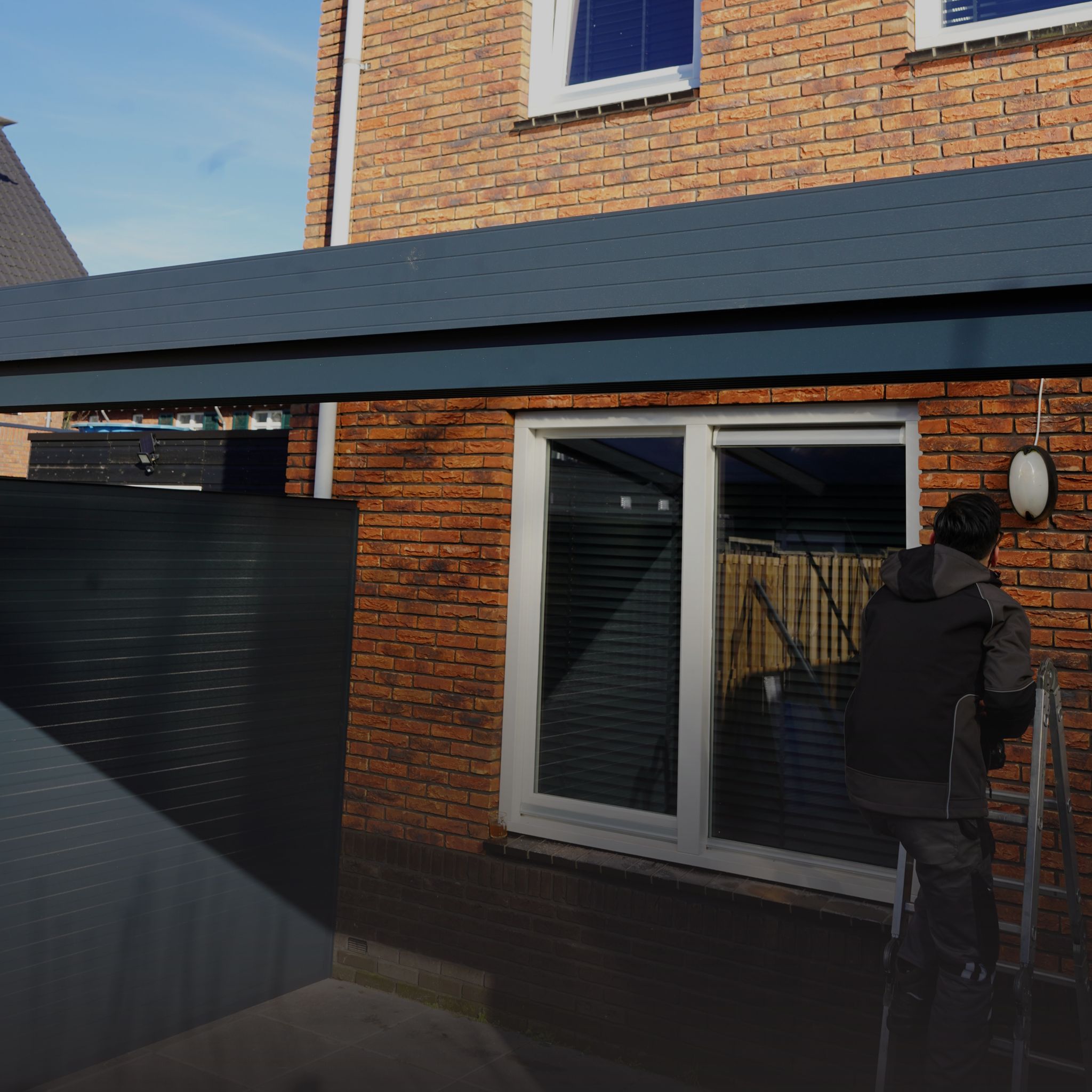 Een man staat op een ladder om een wandlamp te installeren aan de gevel van een moderne woning met een bruine bakstenen muur en een zwarte overkapping.