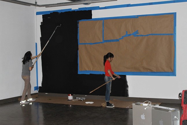 A photo of two girls painting a wall around a certain shape.
