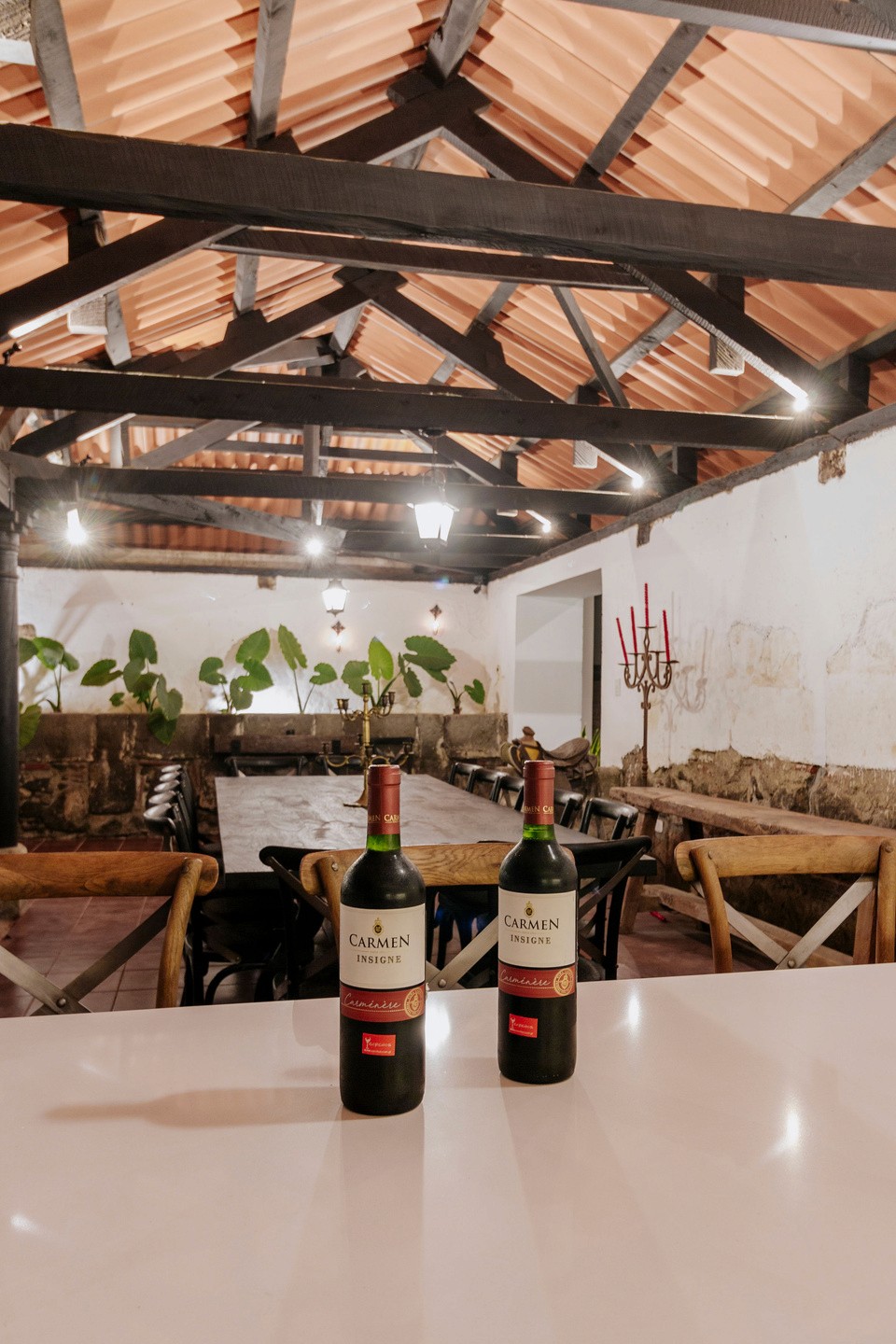 Two bottles of red wine sit on a modern white countertop in a rustic dining area with high wooden beams, a long communal table, and ambient lighting, providing a cozy, inviting atmosphere.