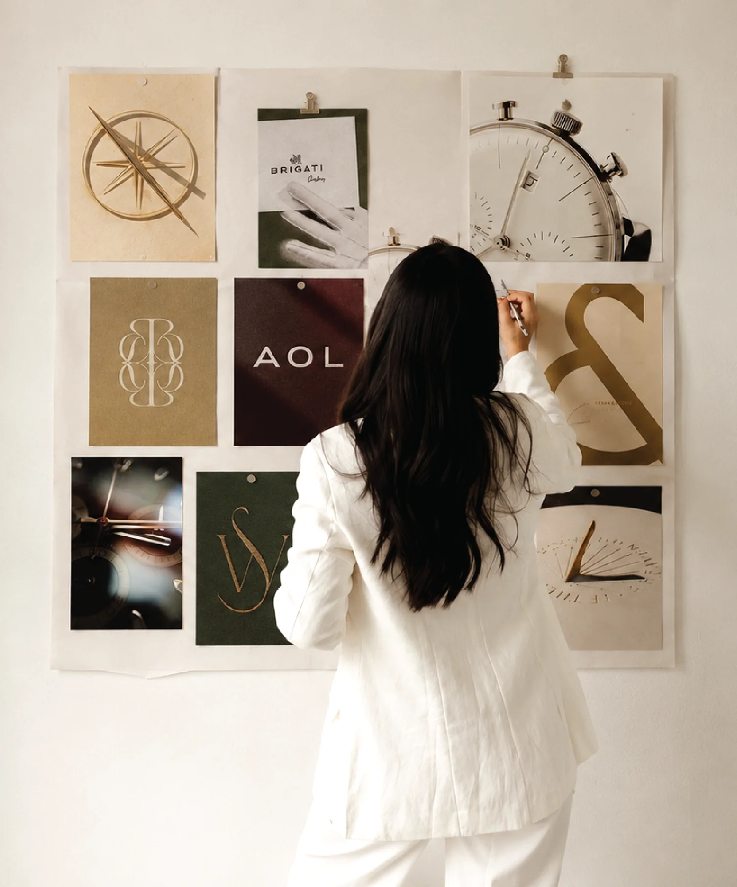 A person in a white suit is pinning a paper with a circular design onto a wall collage featuring various artistic posters, including images of a compass and abstract symbols, creating a visually creative and inspiring workspace.