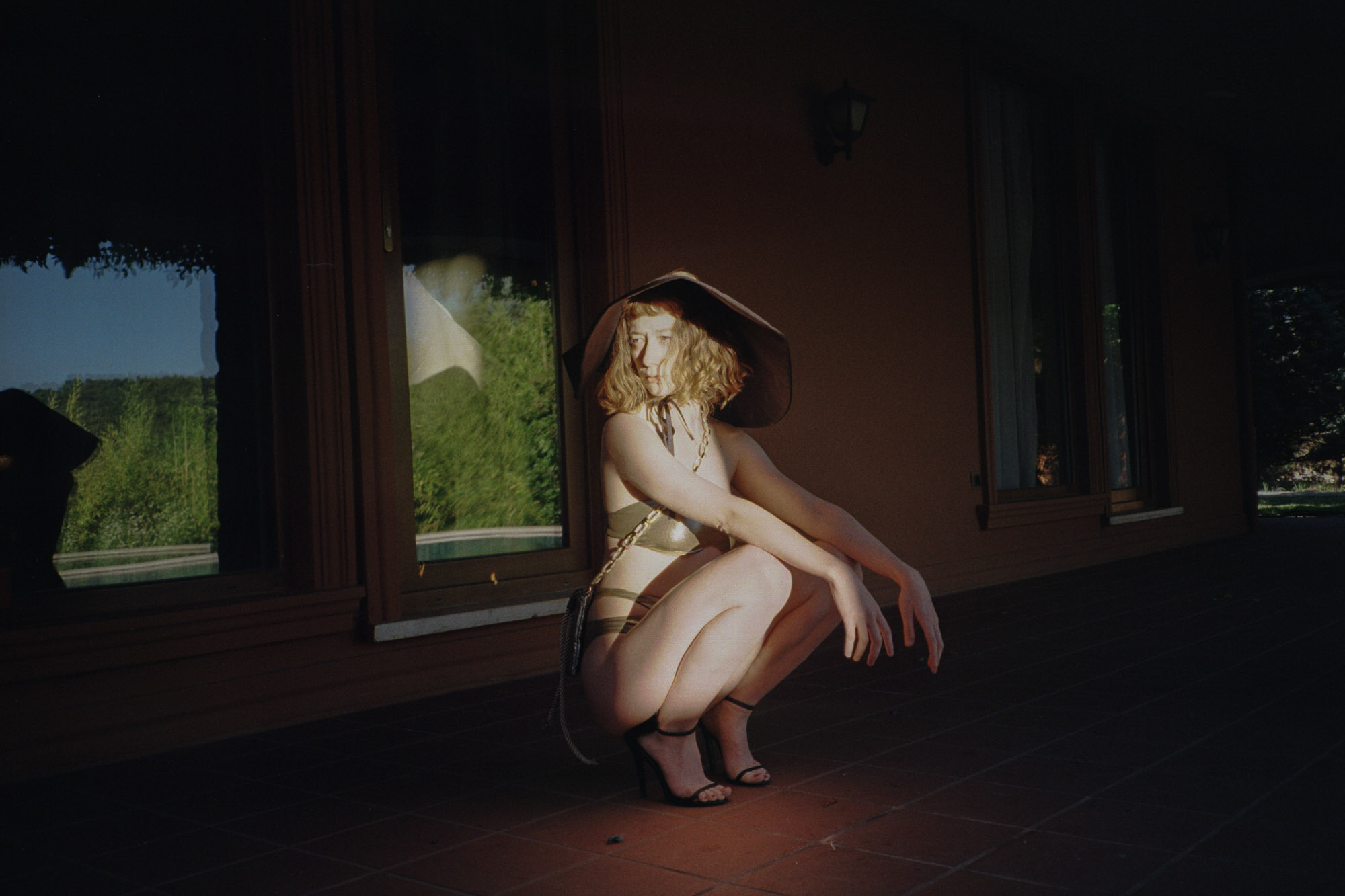 A person in a metallic, space-inspired outfit and large hat poses dramatically on a tiled patio, casting a distinct shadow against the backdrop of a dimly lit building with large windows.