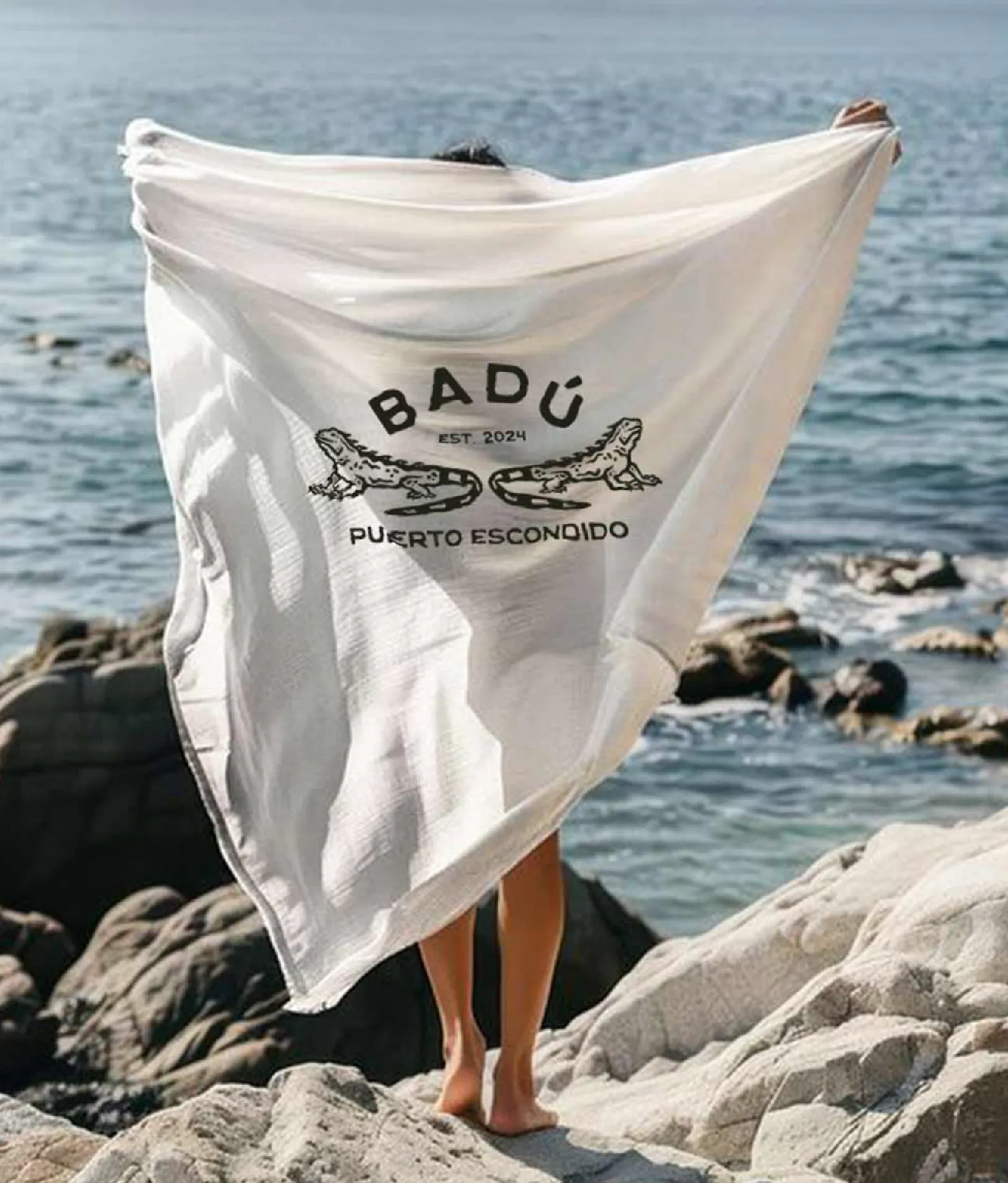 Badu logo with 2 iguanas printed on a beach towel, held up by someone on the beach