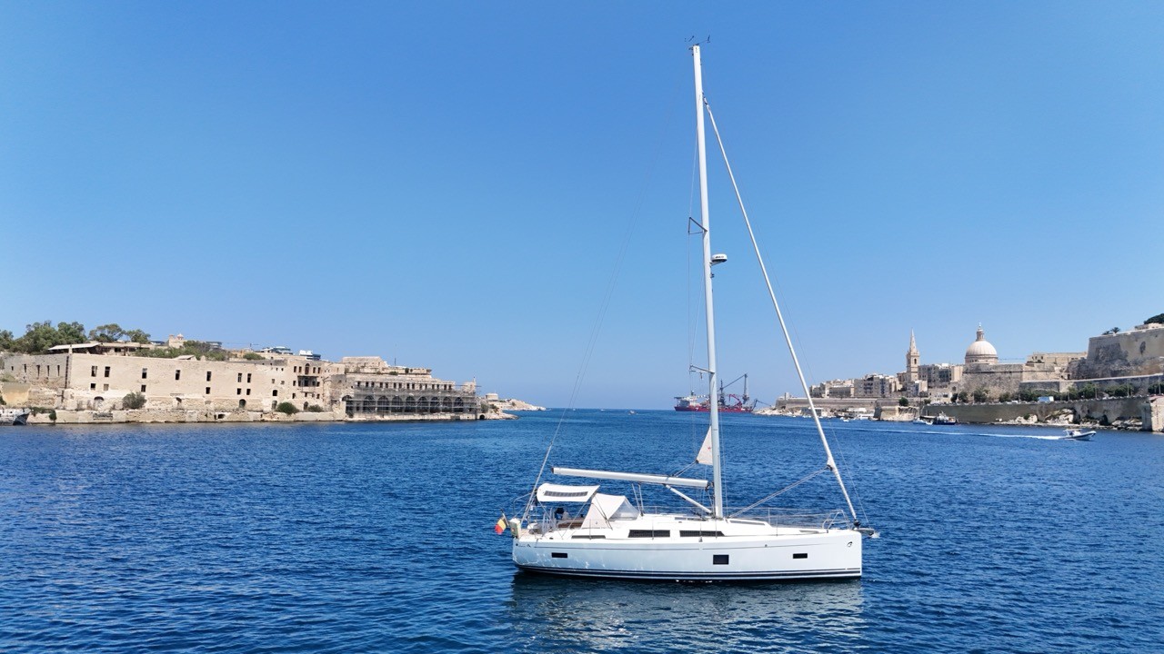A Hanse 388 sailing yacht anchored in a harbor channel between two historic fortified peninsulas, with a domed church and city skyline visible on the far shore under a clear blue sky.