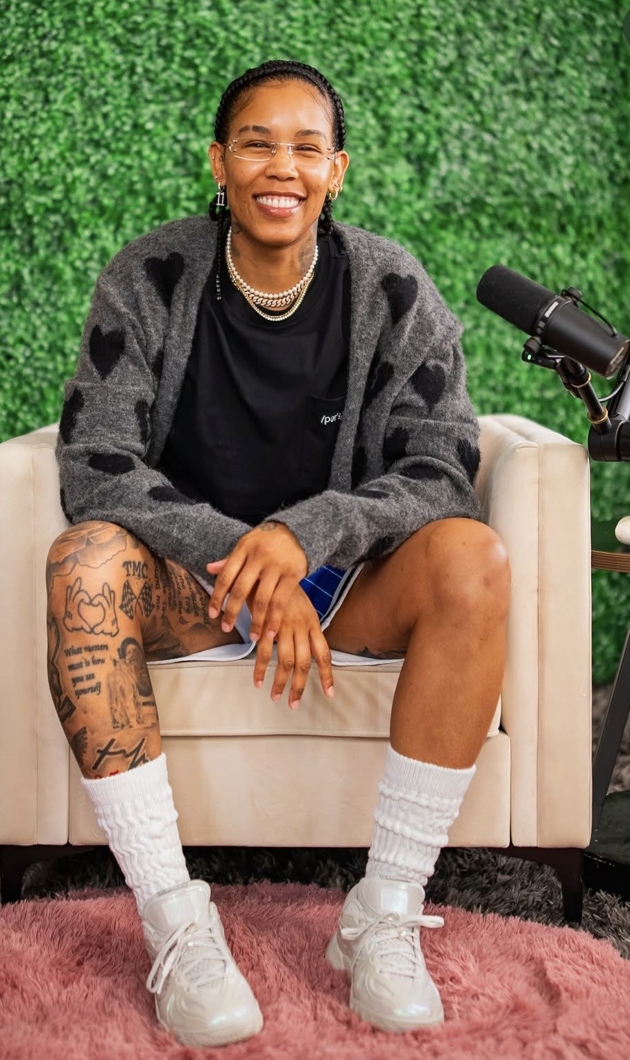 Ty Young with glasses and braided hair sits smiling on a light chair. They wear a gray heart-patterned cardigan, black shirt, shorts, white socks, and sneakers. Visible leg tattoos. A microphone is on a stand nearby, and theres a green leafy background.