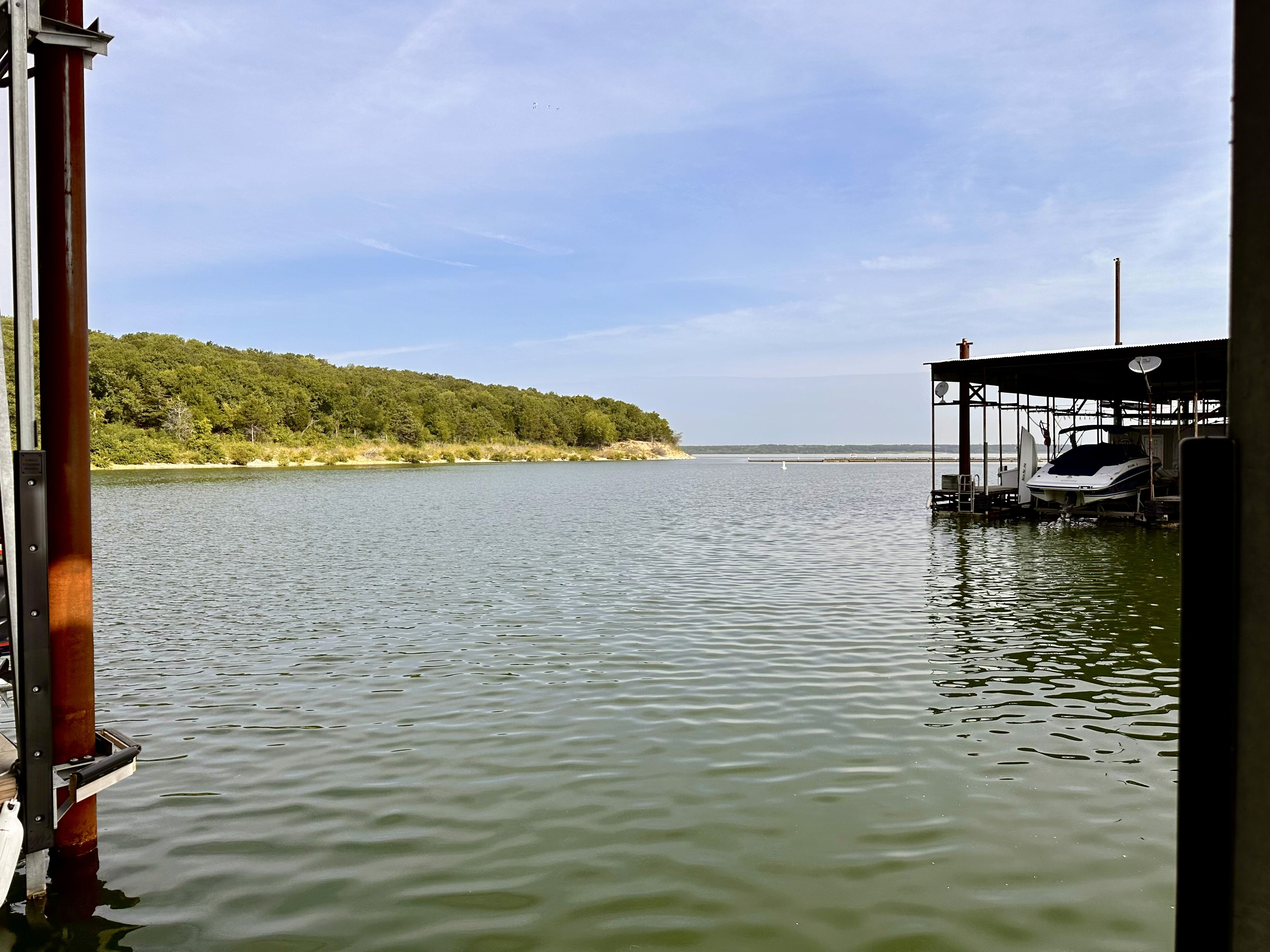 A serene lake scene featuring calm waters rippling gently beneath a bright blue sky, with lush green trees lining the distant shoreline and a boat moored under a covered dock on the right.