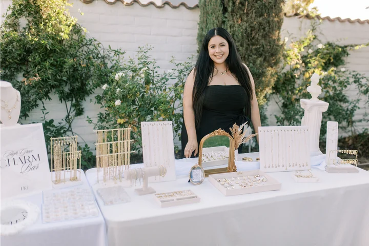 Vendor standing behind a white display table of gold jewelry stands and trays, with a Charm Bar sign, in a garden with a white wall and greenery.