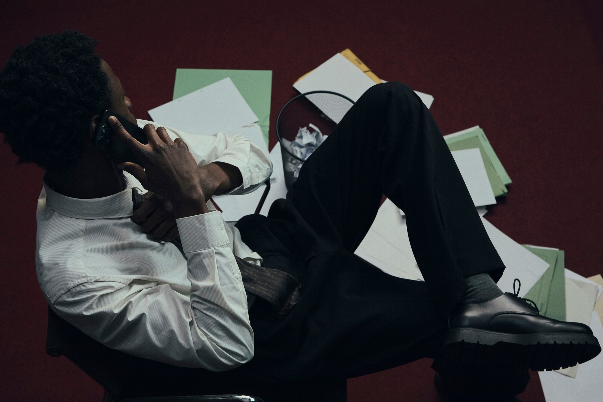 Man in a white shirt reclines in a chair on a red carpet, talking on the phone. Scattered papers surround him, conveying stress or disarray.