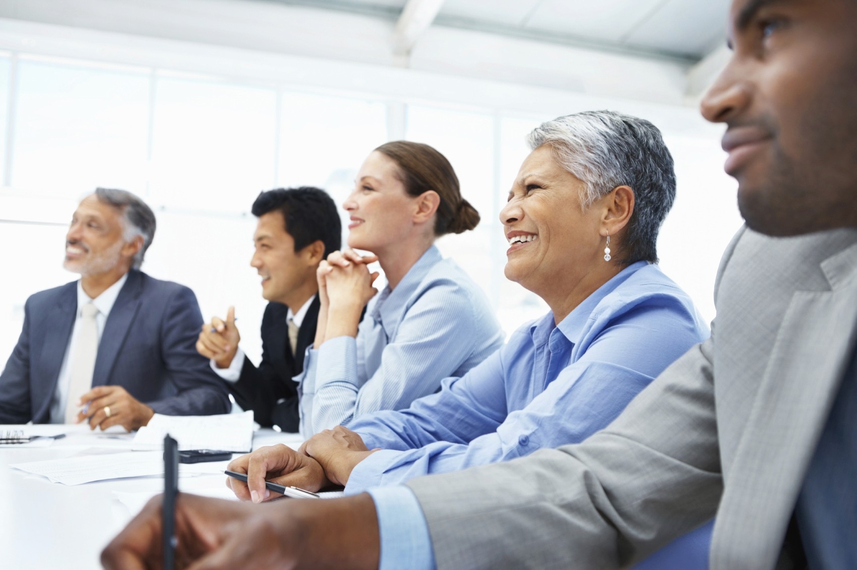 Group of professionals engaged in a meeting, sitting at a conference desk. Smiling faces suggest an exciting discussion. Diverse group in professional attire, focused on something off-camera.