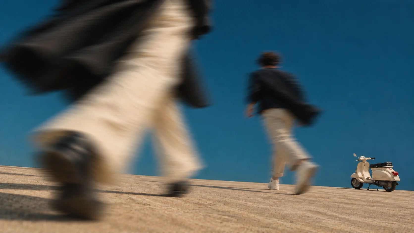 Motion-blurred pedestrians in light trousers and dark jackets crossing a sunlit plain towards a cream vintage scooter