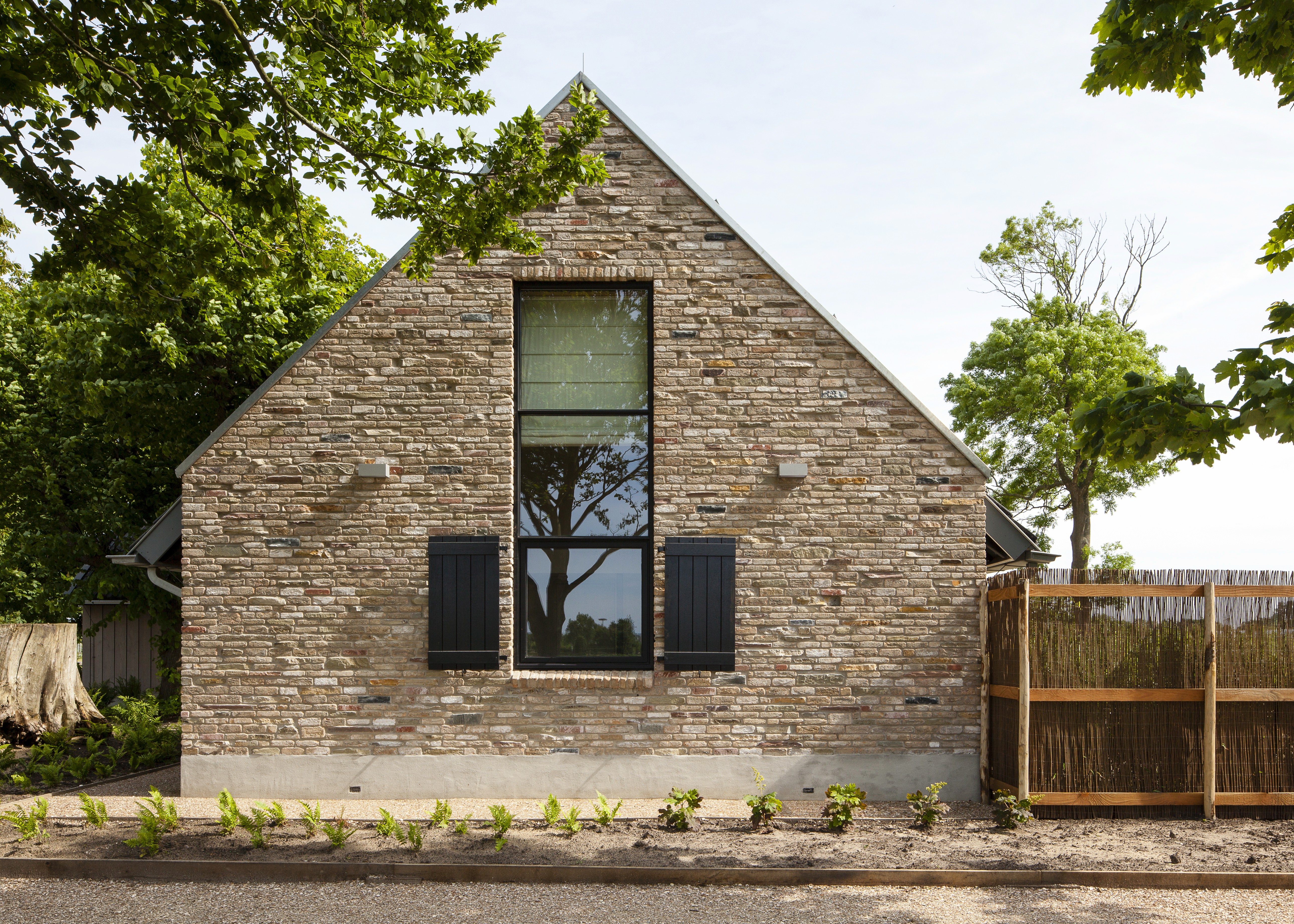 Stenen huis met puntgevel, groot raam en zwarte luiken omringd door groen.