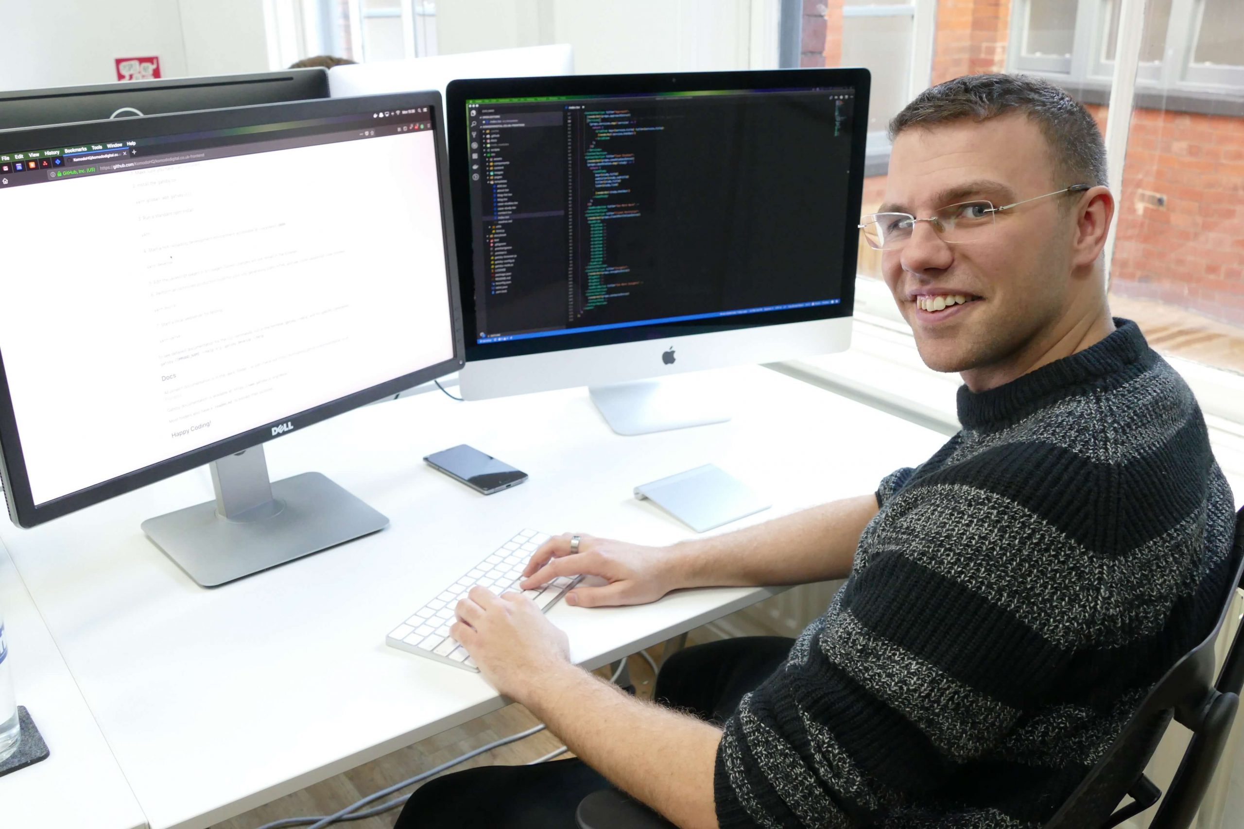 Ian, sat at his desk smiling. There are two parge monitors and a keyboard on the desk