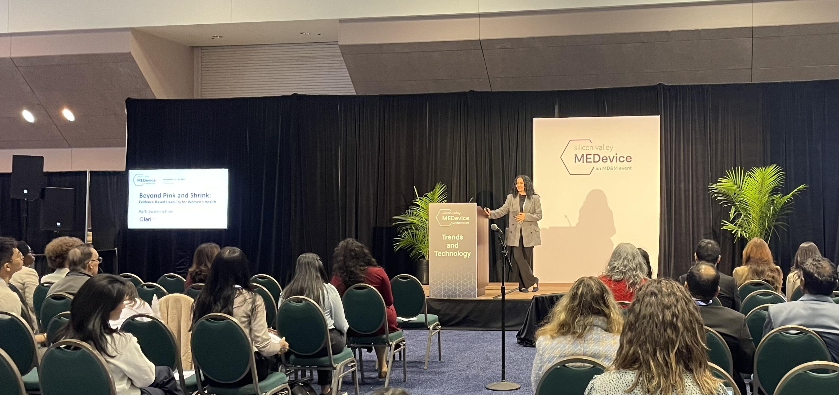 Aarti Swaminathan presenting "Beyond Pink and Shrink: Evidence-Based Usability for Women's Health" on stage at MEDevice Silicon Valley, with audience members seated in the foreground and presentation slide visible on screen.