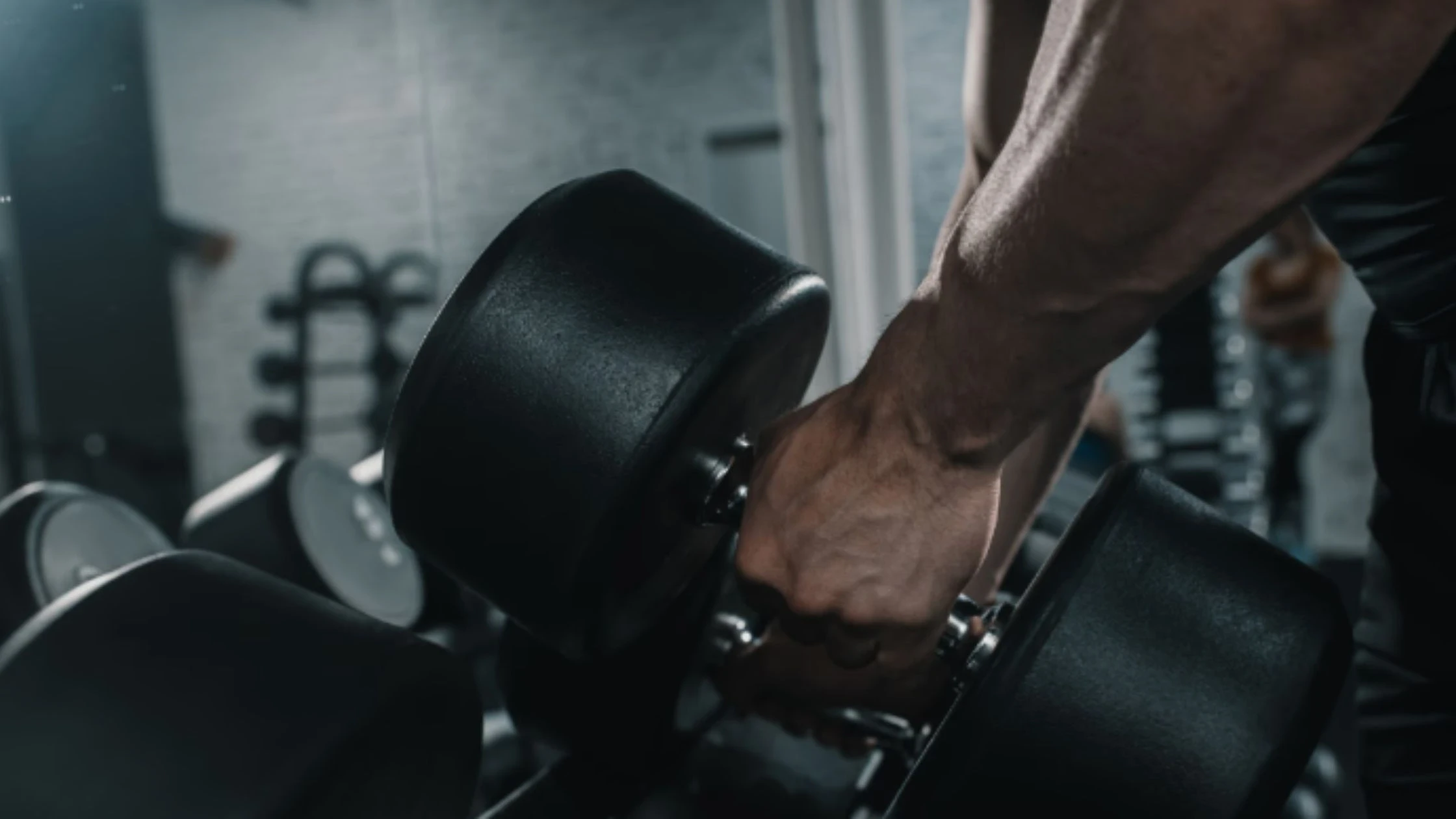 A close-up shot of a person's hand gripping a heavy dumbbell, ready to lift it from a rack in a gym setting. The individual's arm muscles and veins are prominent, indicating the effort required to lift the weight. The gym background is out of focus, with some visible weights and equipment in the distance. The lighting is dim, creating a focused and intense atmosphere that emphasizes the strength and effort involved in the workout.