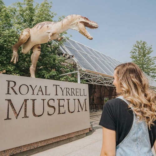 Person in overalls stands facing a Royal Tyrrell Museum sign with a dinosaur sculpture above it. Trees and a glass-roofed structure are visible.