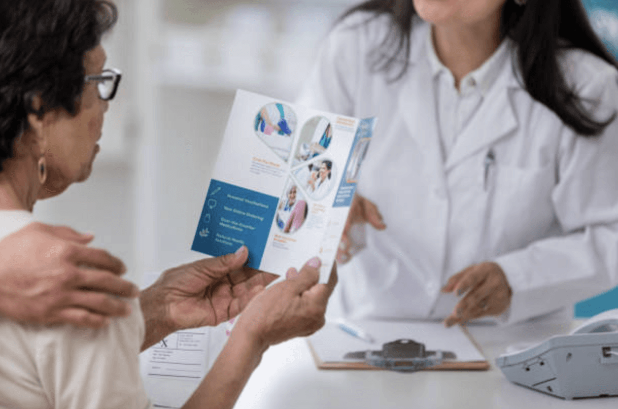 Older woman reading a white and blue pamphlet. In front of her is a female doctor in a white lab coat.