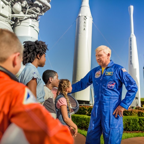 En man i en blå NASA-rumddräkt pratar med en grupp barn med raketer i bakgrunden på en solig dag.