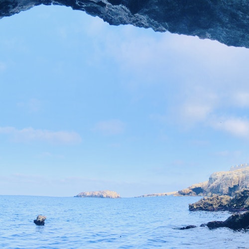 View from inside a cave looking out to a calm sea, with rocky cliffs and small islands in the distance under a clear blue sky.