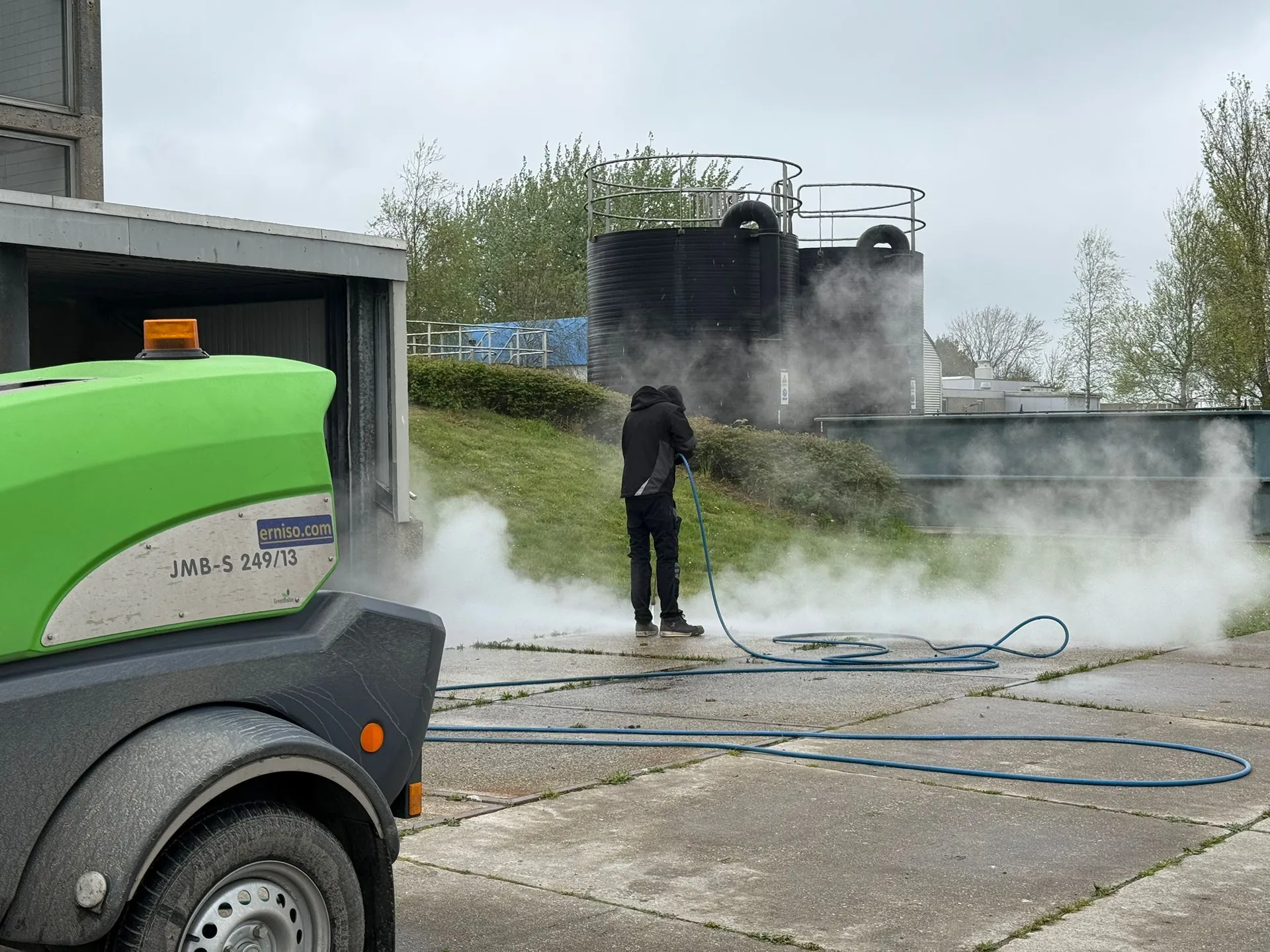 Medewerker reinigt een betonnen buitenverharding met een heetwaterhogedrukreiniger; de stoom slaat van het beton af en links staat de groene aggregaatwagen met de slang