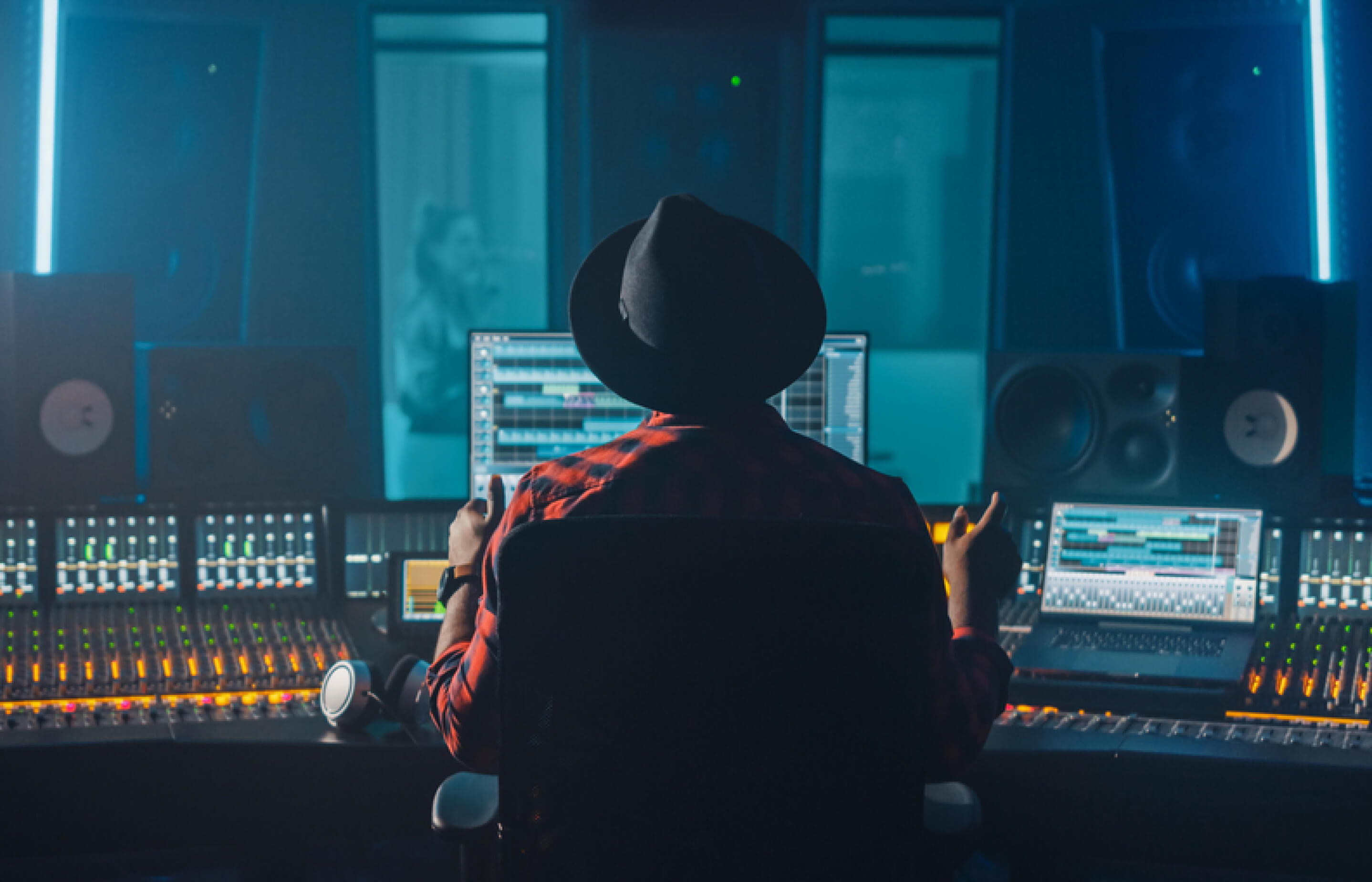 A person in a black hat sits at a desk, focused on multiple screens in a dimly lit sound studio.
