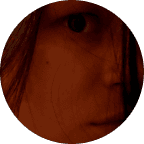 Blonde woman in a black long-sleeve and apron, arms crossed—minimalist studio portrait on a beige background.