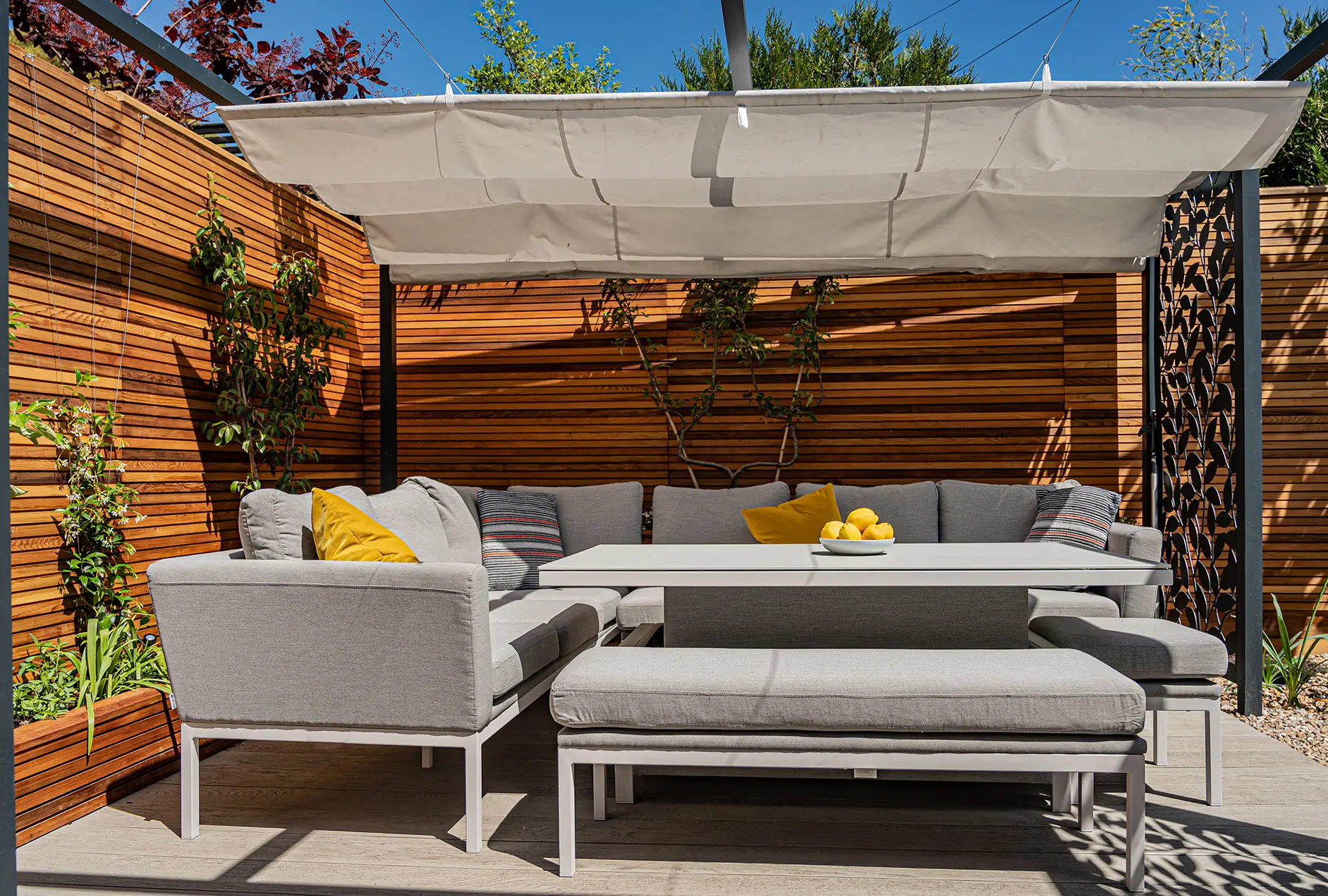 A cozy outdoor seating area with gray sofas and yellow cushions, surrounded by wooden fences and greenery.
