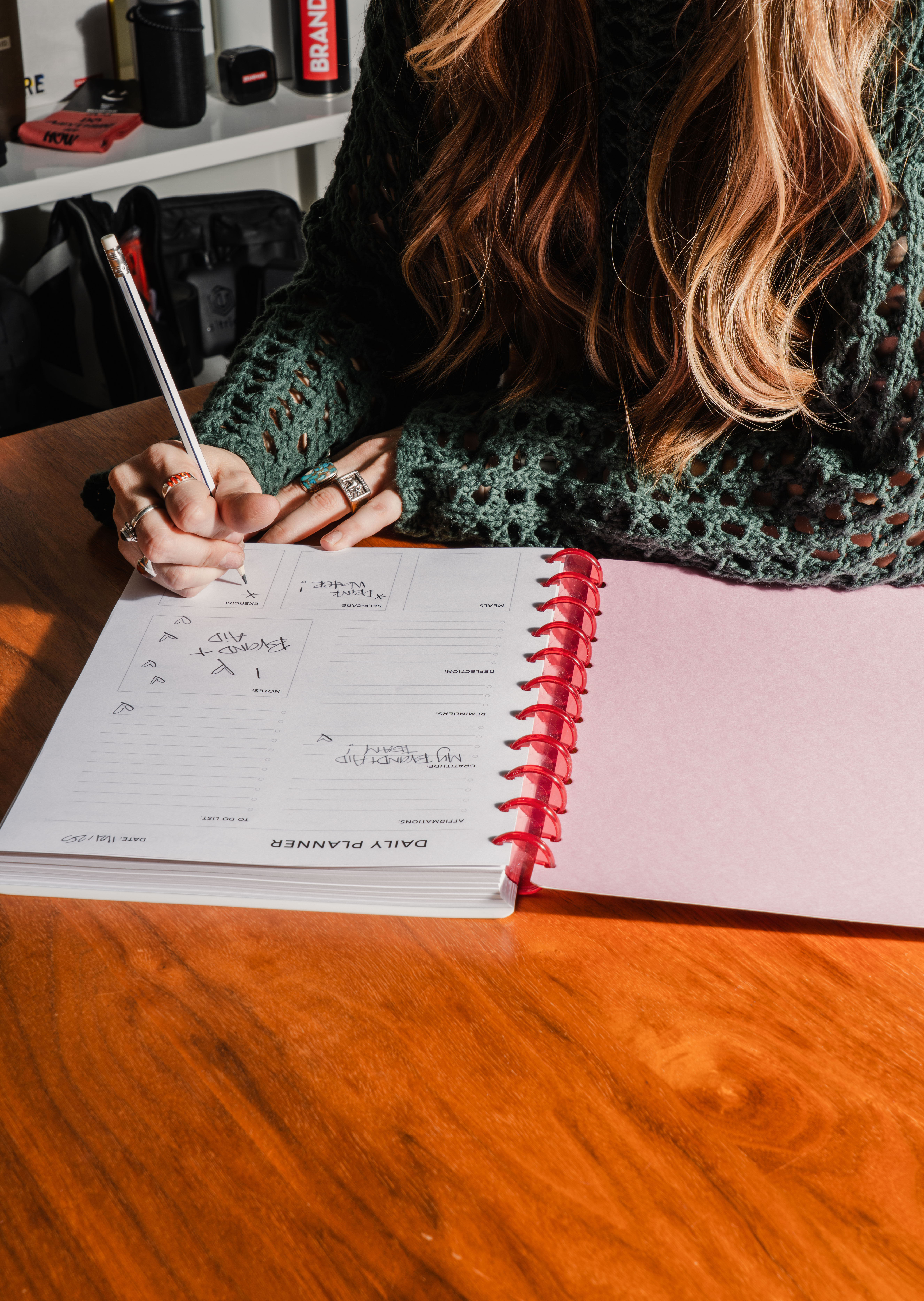 Girl in a green sweater writing in an open daily journal planner with a pink front page and binding on a wood table.