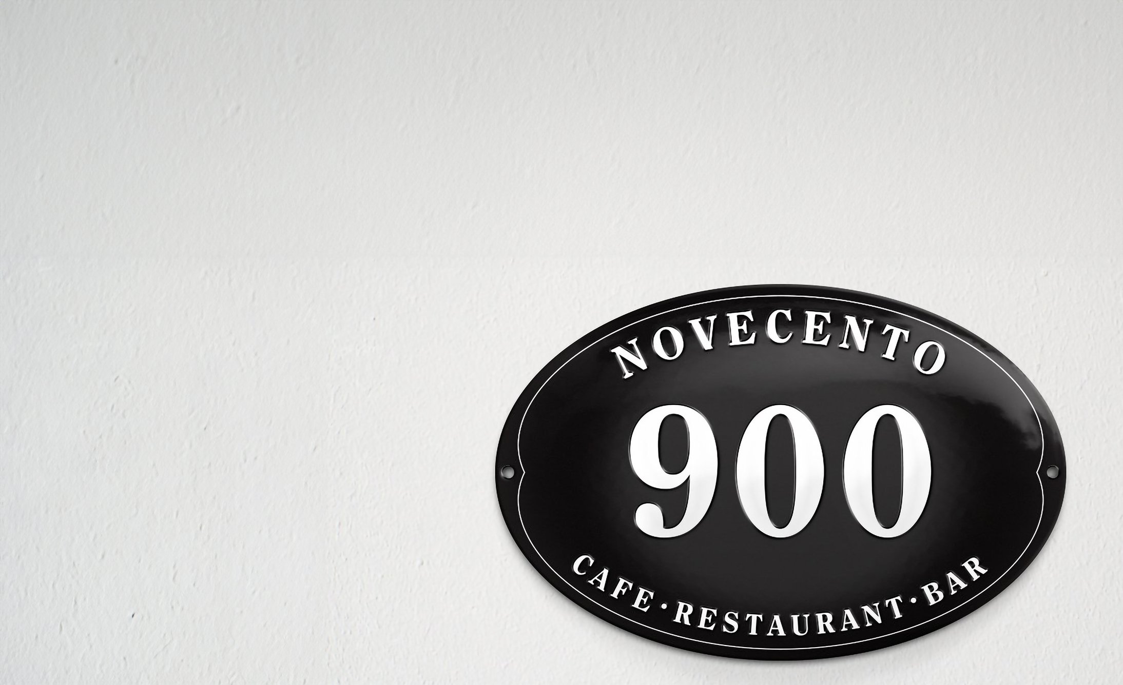Oval, black sign on white wall with "Novecento 900 Cafe Restaurant Bar" in white, bold letters. Minimalist design conveys a modern, sophisticated tone.