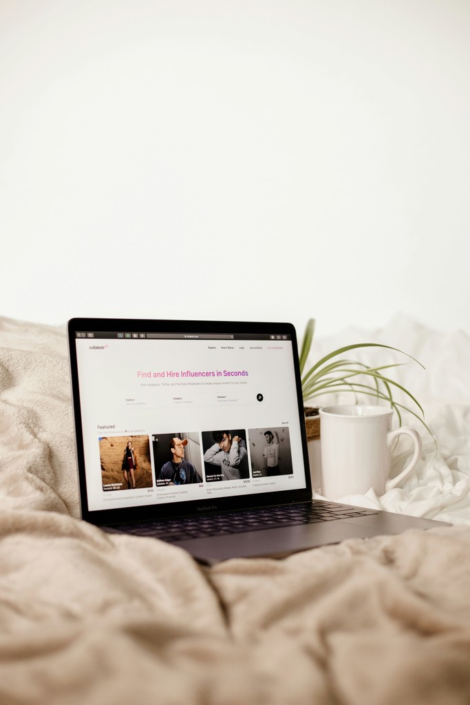 A cozy scene features a laptop displaying an influencer hiring platform on a bed, accompanied by a coffee mug and a plant, symbolizing the convenience and comfort of remote work and digital marketing.