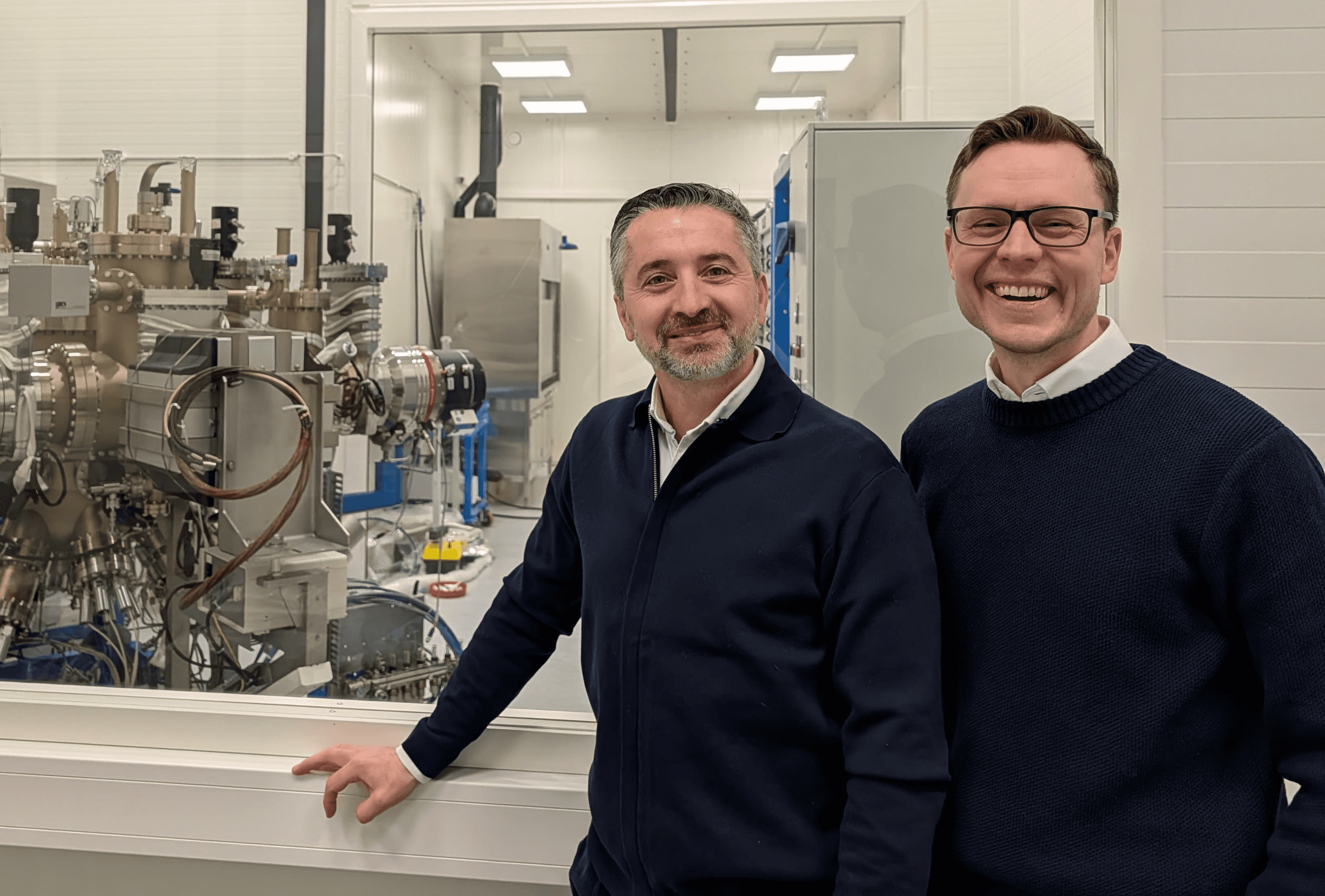 Mircea Guina and Jussi-Pekka Penttinen, founders of Vexlum, smiling in front of advanced laser technology equipment inside a high-tech laboratory.