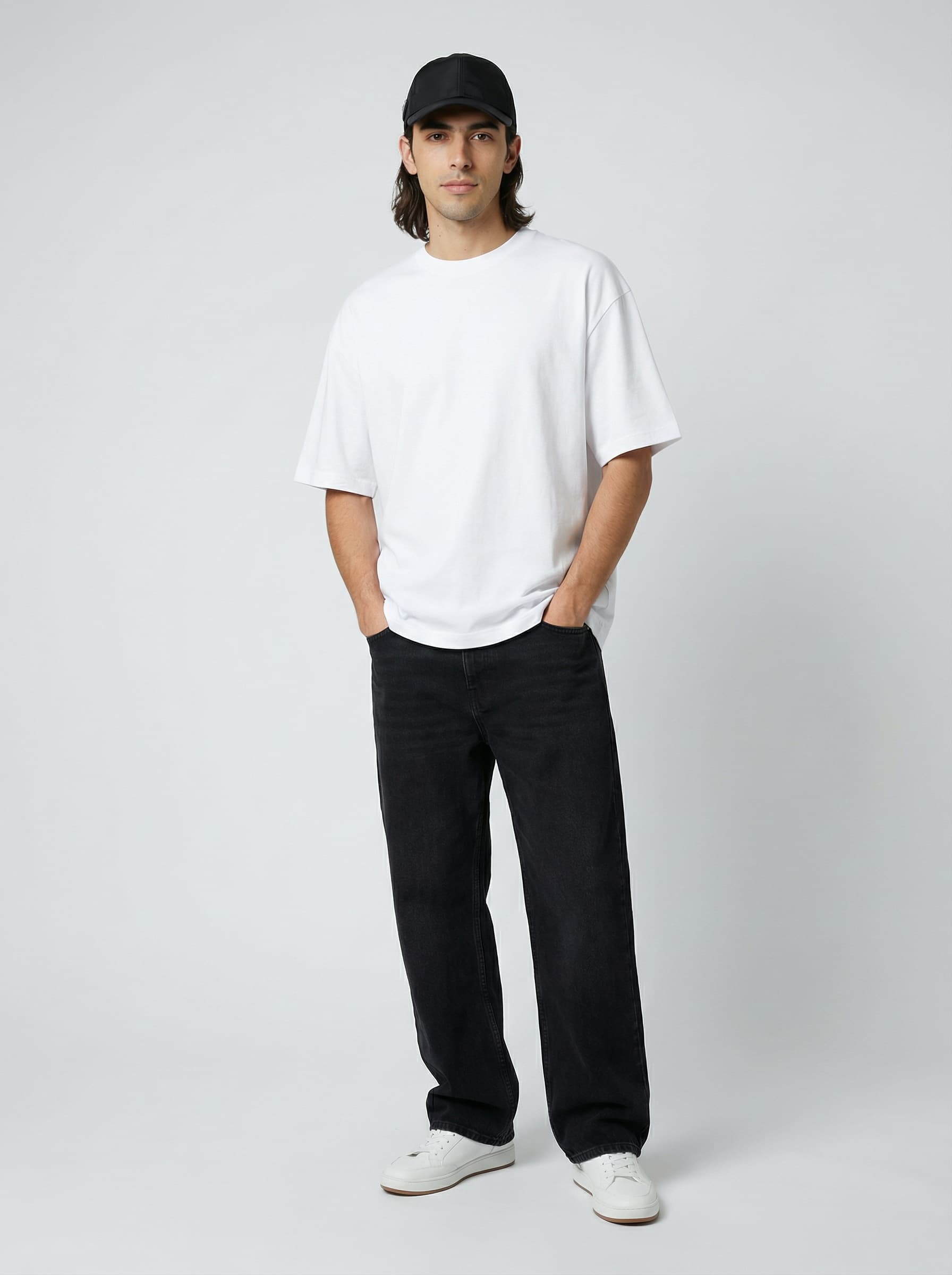 A person stands against a plain white background wearing a black baseball cap, a white oversized T-shirt, black pants, and white sneakers, exuding a casual fashion style.