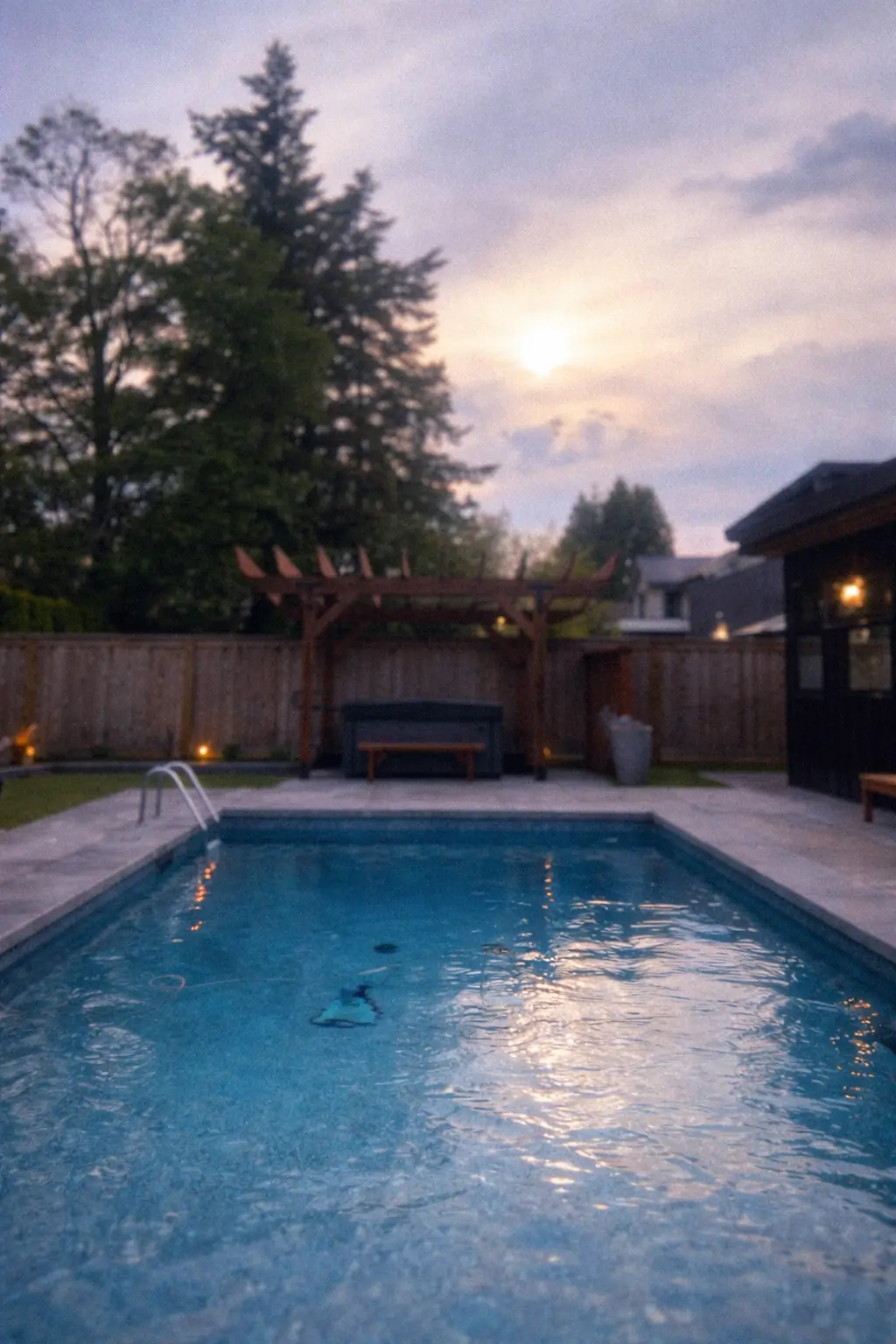 Modern backyard swimming pool with wooden pergola seating area and privacy fence at sunset.