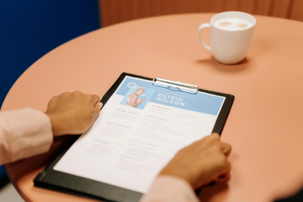 A person reviews a resume labeled "Olivia Wilson" on a clipboard placed on a round, light brown table beside a cup of coffee.