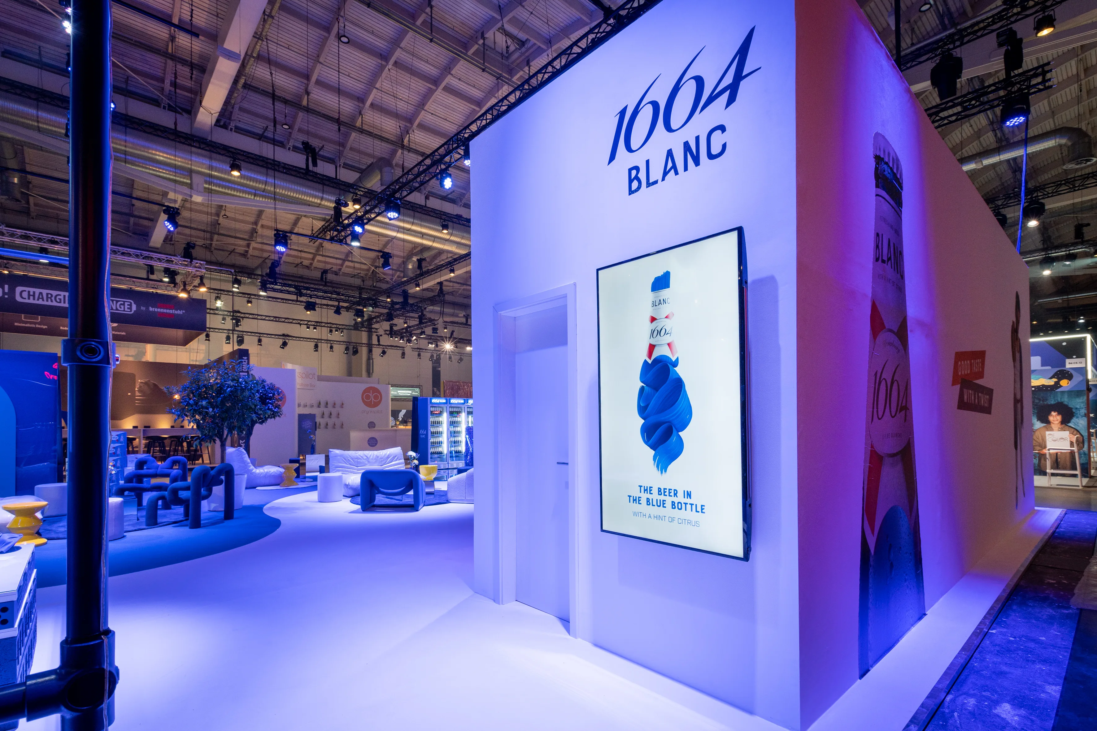 White outer wall of the 1664 Blanc booth at OMR with logo, screen showing the blue bottle and violet-lit lounge behind.