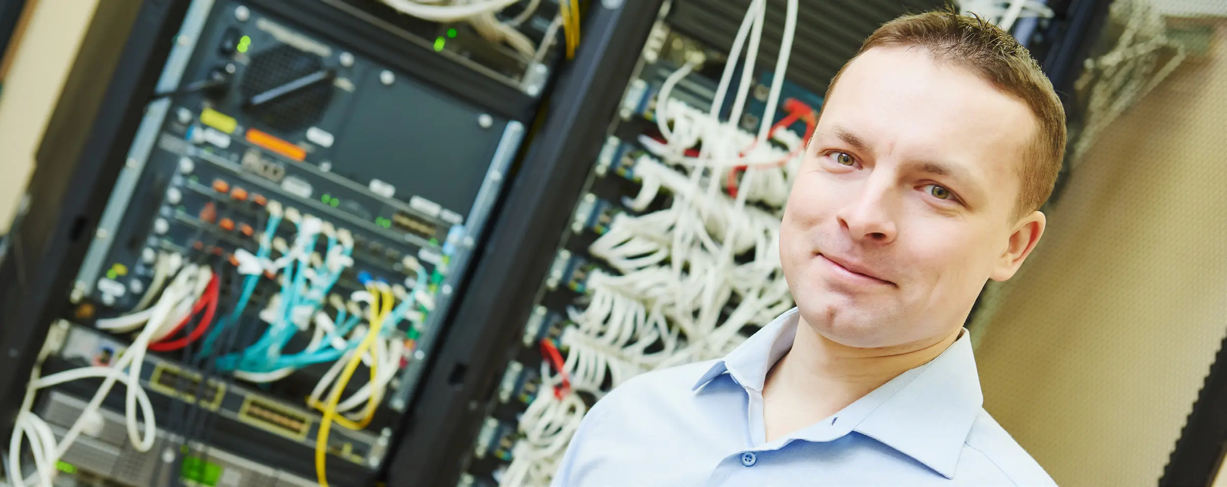 Eine lächelnde Person steht neben einem Serverrack, das mit verschiedenen elektronischen Bauteilen gefüllt ist.