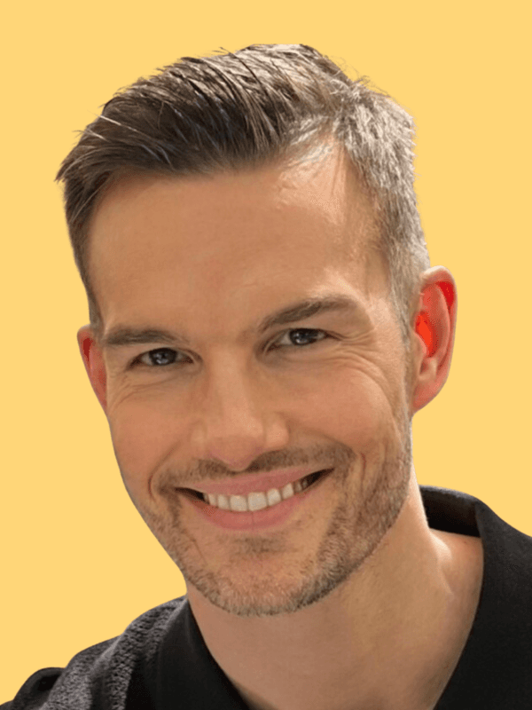 A smiling young man with crossed arms, wearing a plaid shirt and white t-shirt, poses against a dark background.
