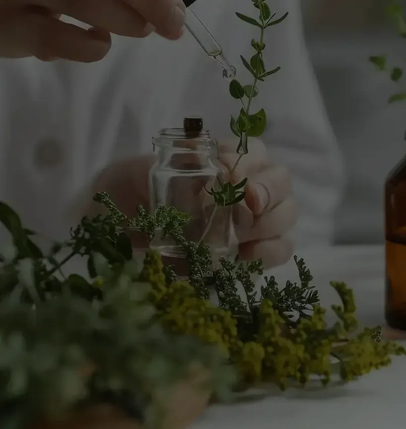 A person in a white lab coat uses a dropper to add liquid to a small glass jar amidst various fresh green herbs and amber essential oil bottles.