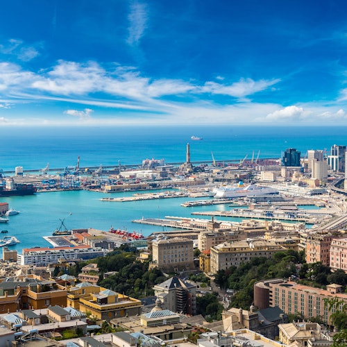Aerial view of a coastal city with a busy port, ships, and buildings under a bright blue sky with scattered clouds.