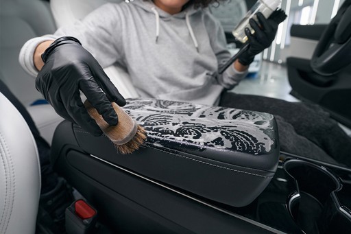 A person wearing black gloves uses a brush to apply a cleaning solution from a spray bottle onto a car's center console, detailing the interior.
