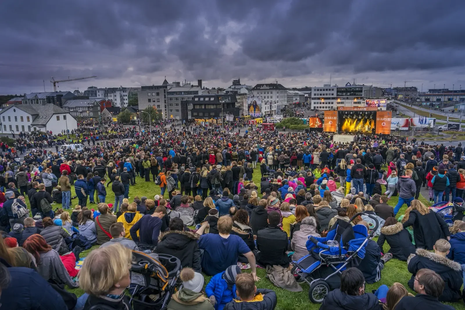 A massive crowd of people sits and stands on a grassy hillside in downtown Reykjavík during the Culture Night outdoor concert. An illuminated stage with yellow lights is visible in the background against a dramatic, overcast evening sky.