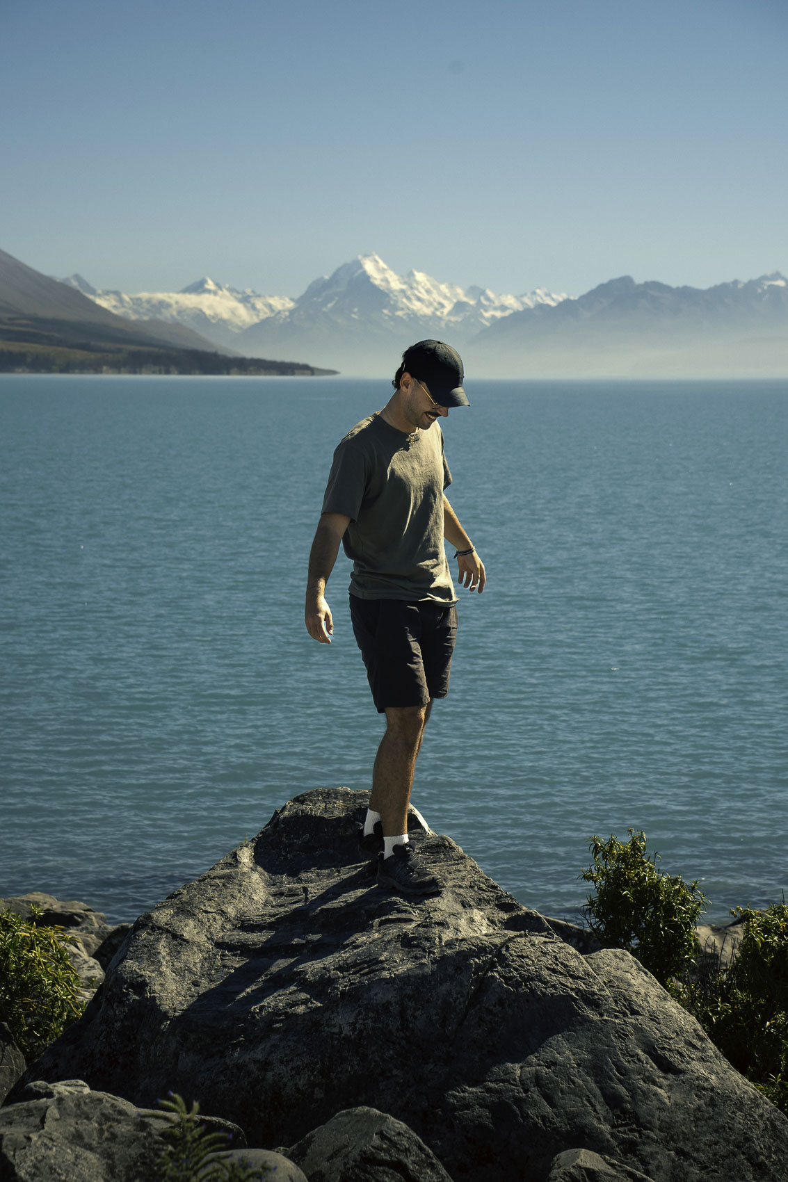Photographe debout sur un rocher au bord d’un lac en Nouvelle-Zélande avec vue sur les montagnes et le Mont Cook, photographie de voyage par Robin Masson