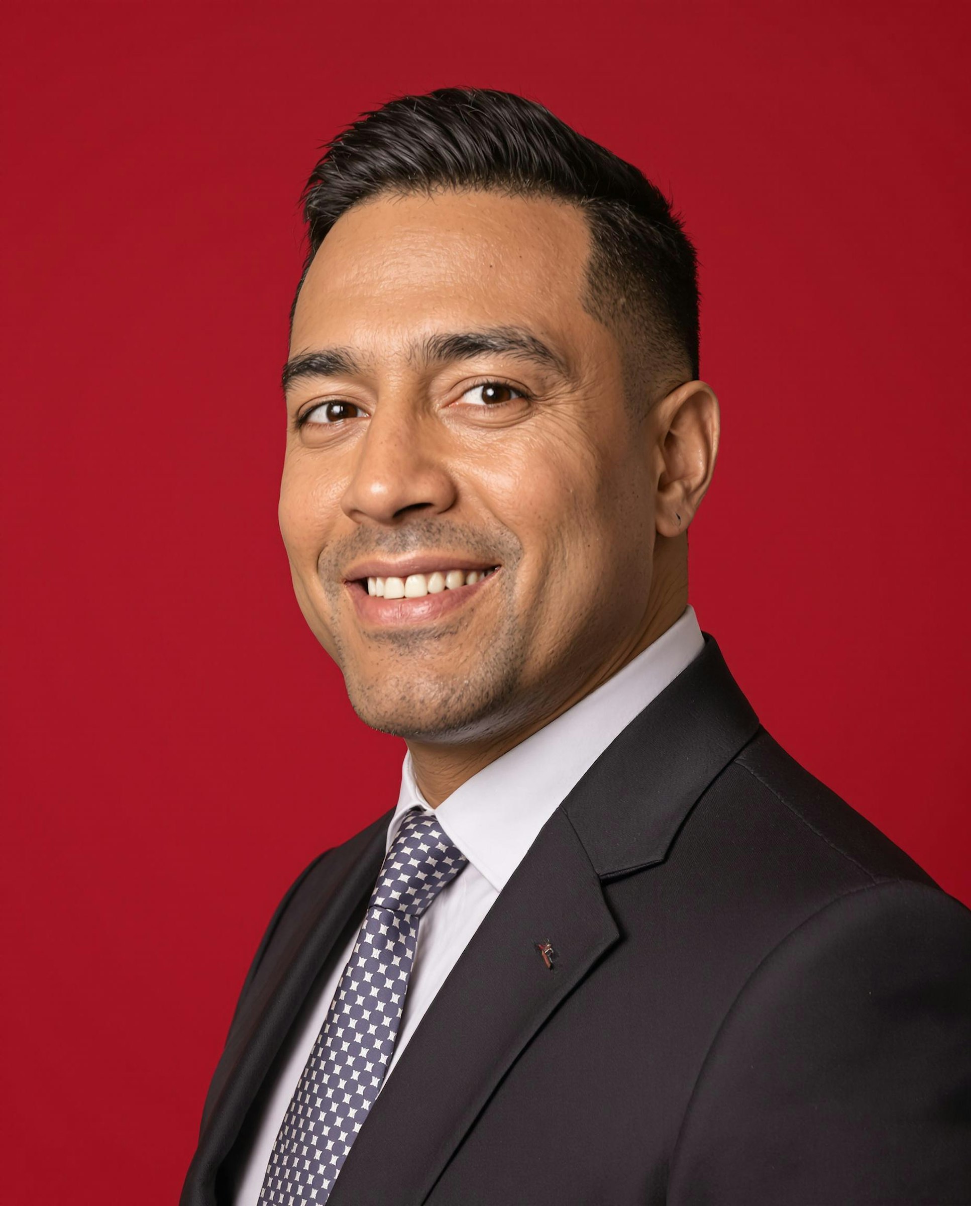 A smiling man in a suit and tie against a bold red background, exuding professionalism and confidence.