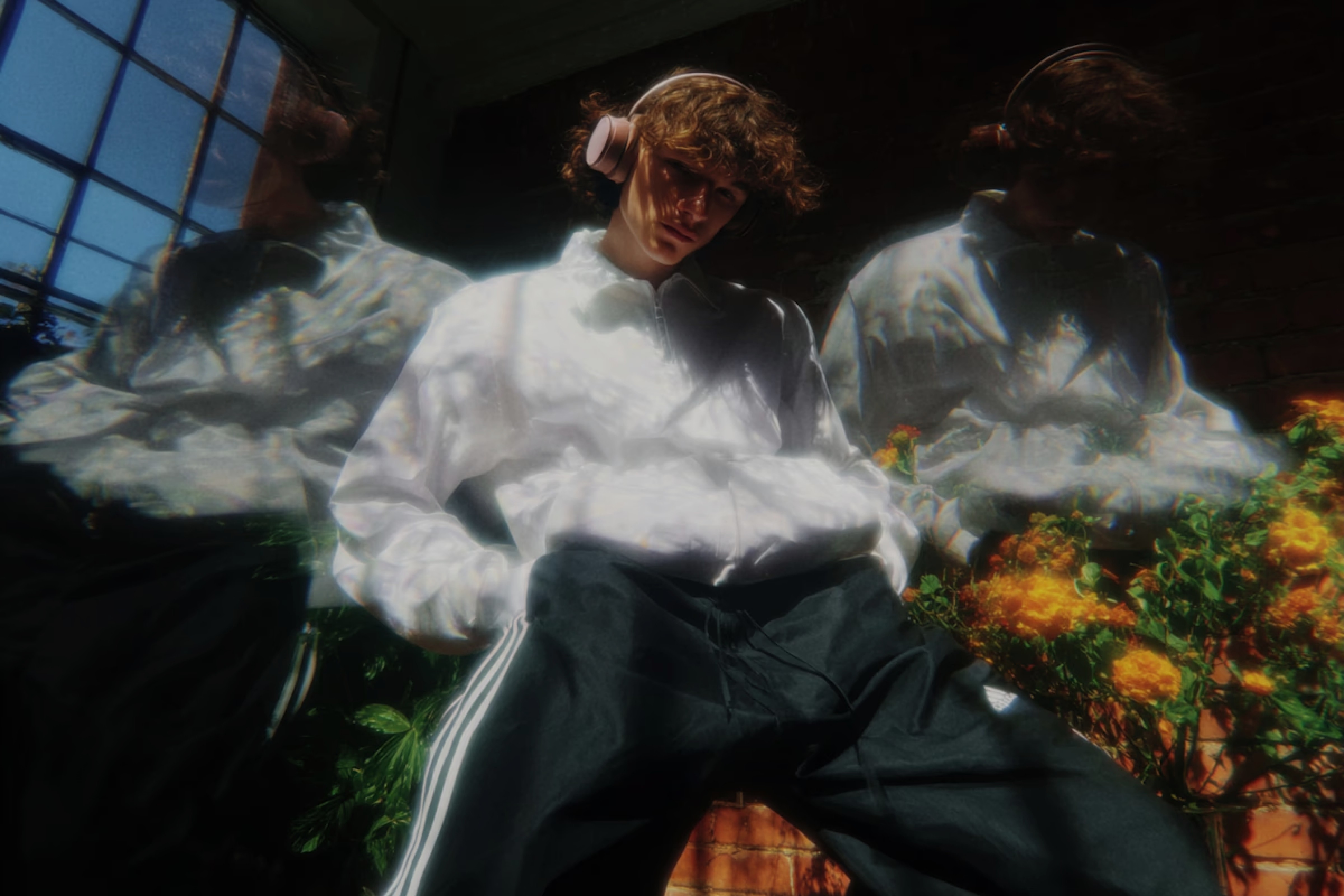 Young man with curly hair and headphones wearing a white jacket, flanked by prismatic ghosting effects and yellow flowers.