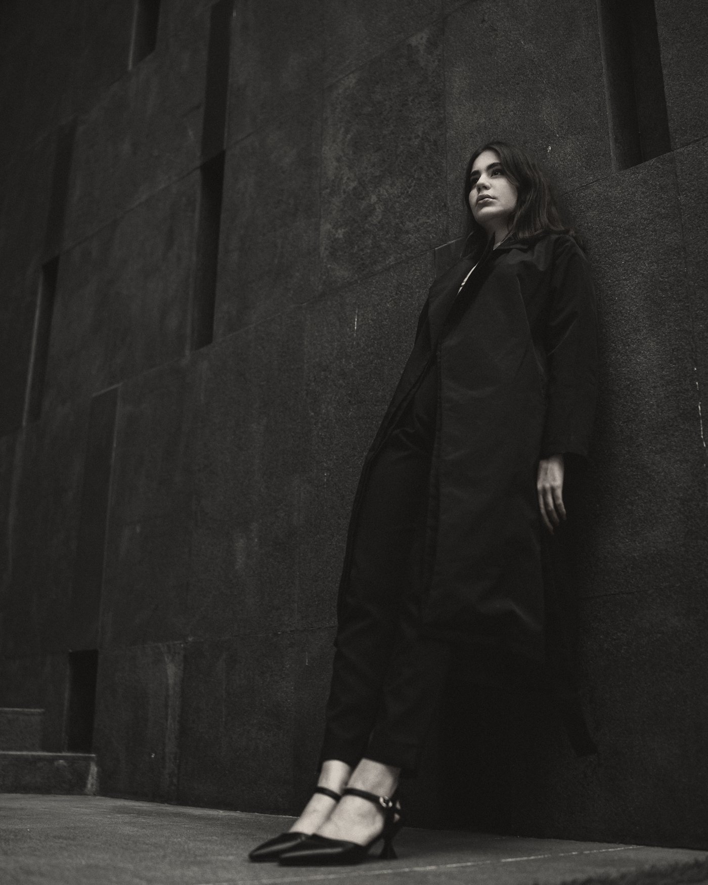 Model leaning on a dark stone wall in a long black coat
