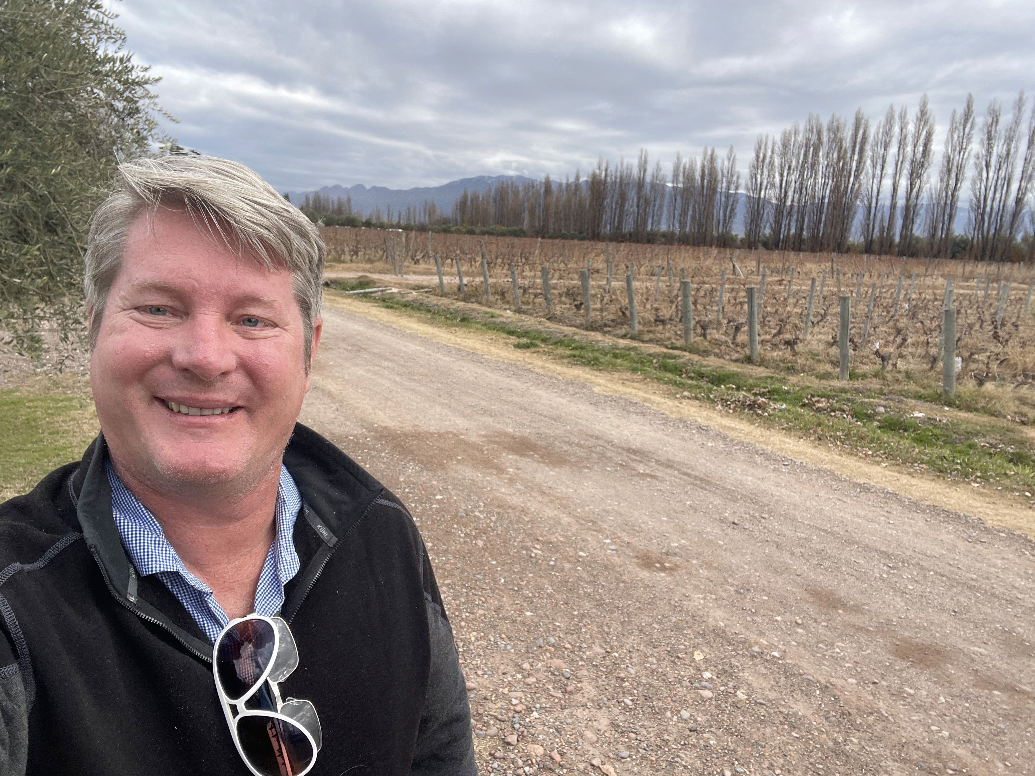 Brian Peddie smiling near a vineyard path with mountains in the distance.