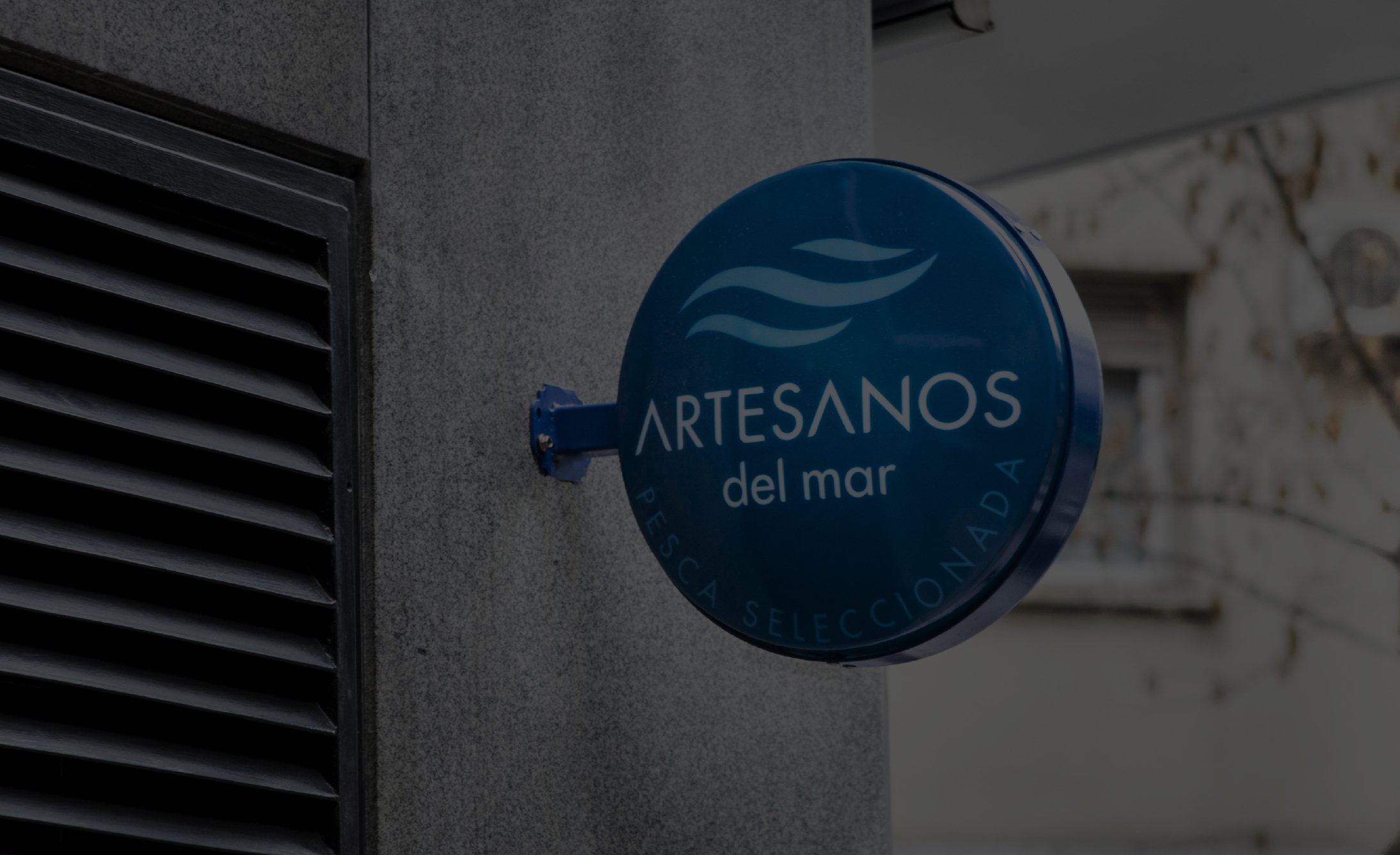Blue circular sign on a gray wall displays "Artesanos del Mar" with wave motifs. Background includes a blurred building, conveying a coastal theme.